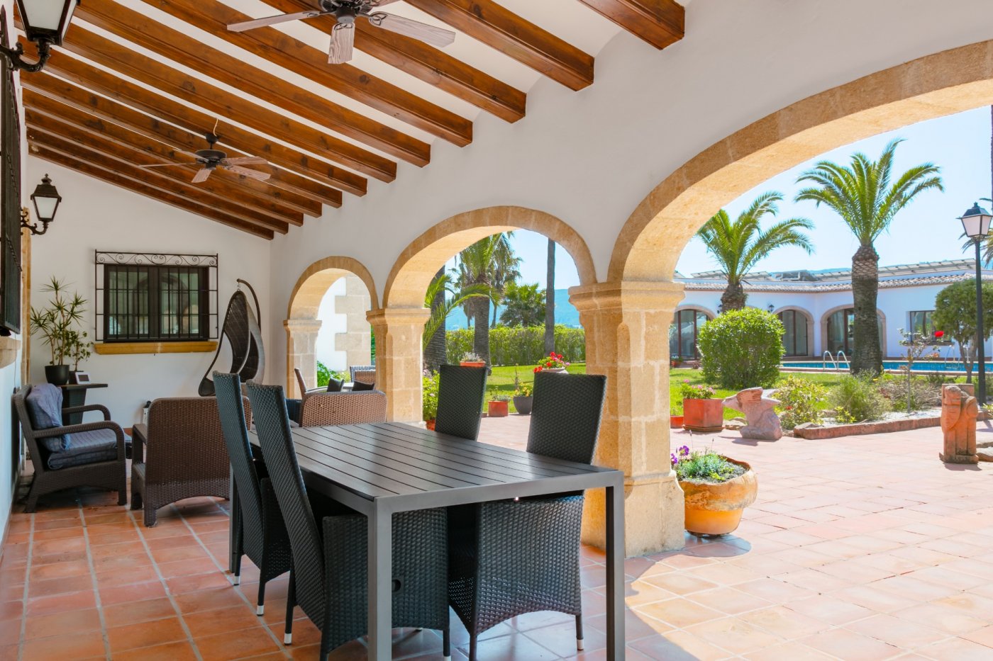 Spacious covered terrace featuring traditional Tosca stone arches, wooden beam ceilings, and a dining area overlooking the palm-filled Mediterranean garden.