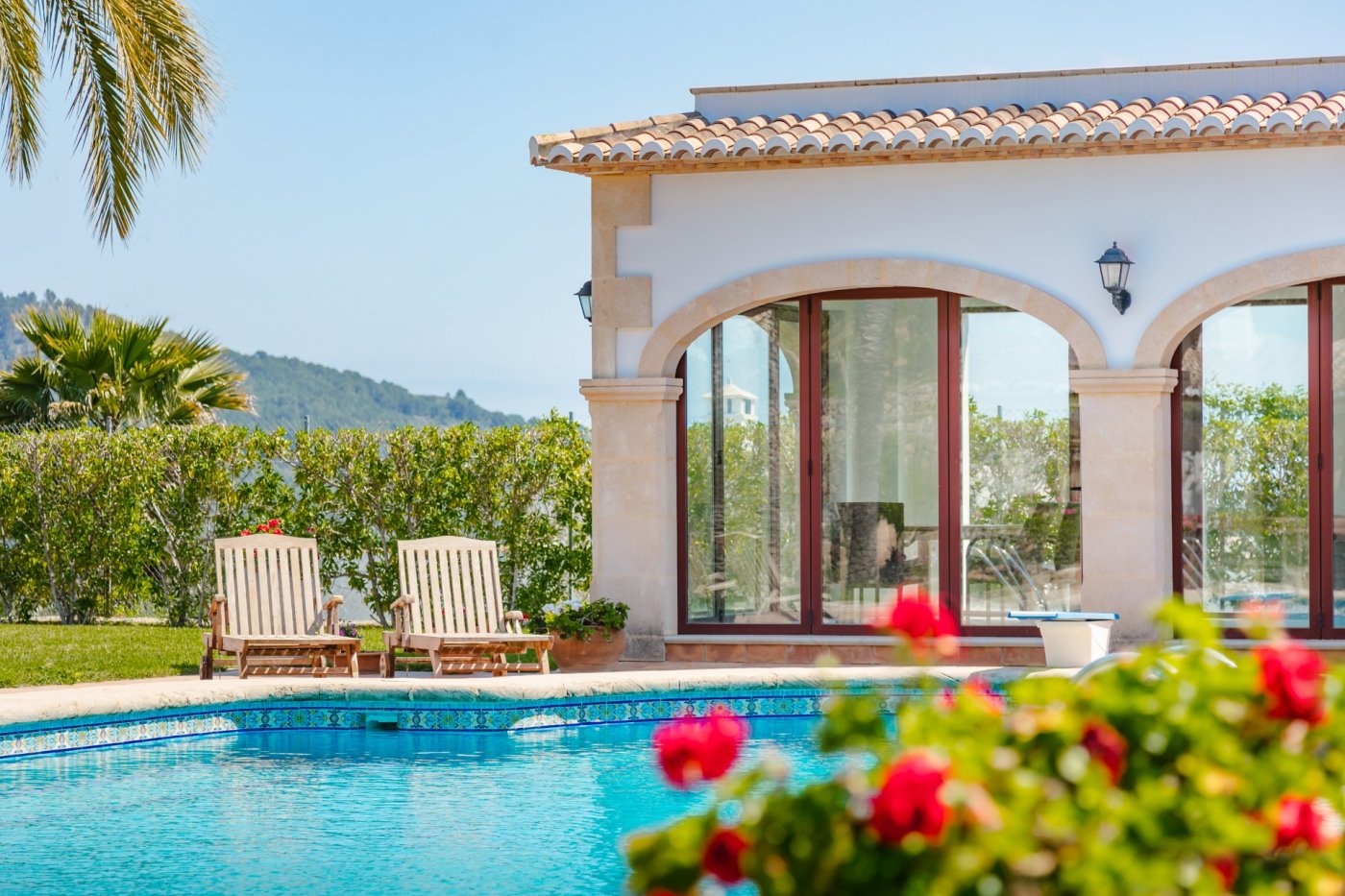 Swimming pool featuring blue mosaic tiling adjacent to a Mediterranean building with Tosca stone arches, wooden window frames, and a sun deck with loungers.