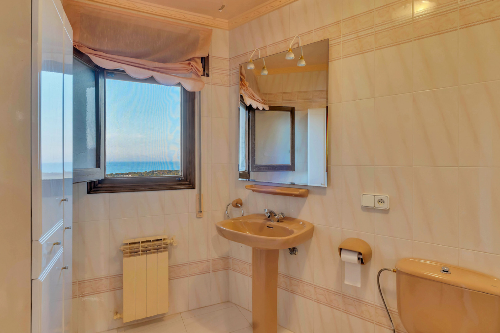 Bathroom featuring a beige pedestal sink, matching toilet, and a window offering clear views of the sea and the surrounding coastline.