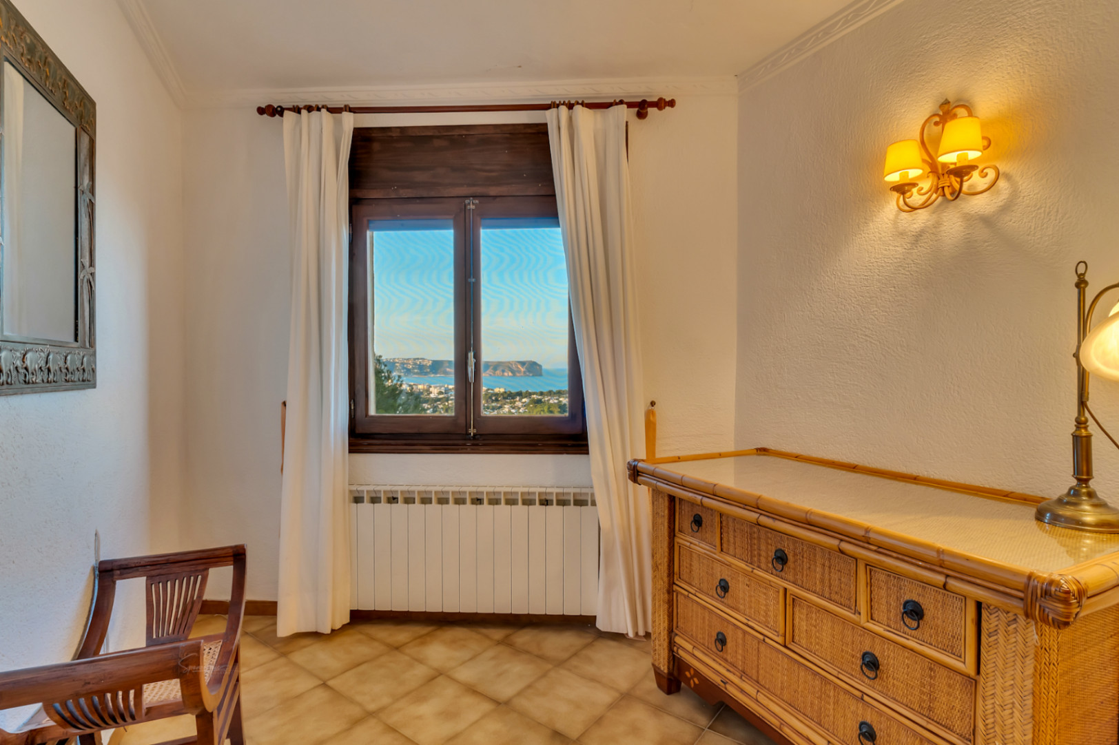 Bedroom corner featuring a wicker dresser, wooden chair, and window overlooking the sea and Cabo San Antonio. Includes a radiator and warm wall lighting.