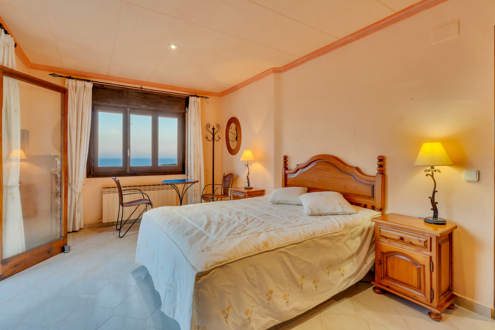 Bedroom featuring classic wooden furniture, hexagonal floor tiles, and a window overlooking the sea. Includes a radiator and a small seating area with natural light.