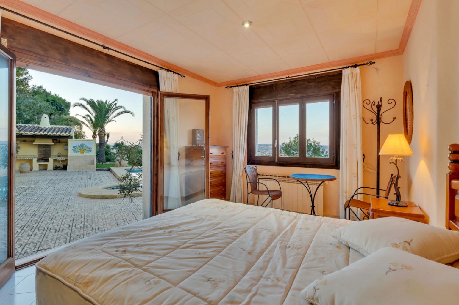 Bedroom featuring dark wood frames and direct access to a paved terrace with views of the pool, palm trees, and outdoor kitchen area.