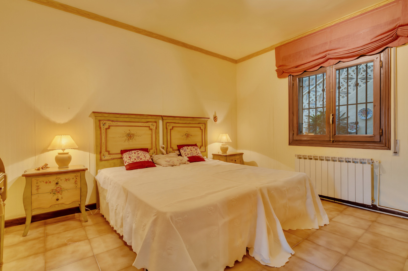Bedroom featuring twin beds with hand-painted wooden headboards, terracotta tile flooring, central heating, and a wood-framed window.