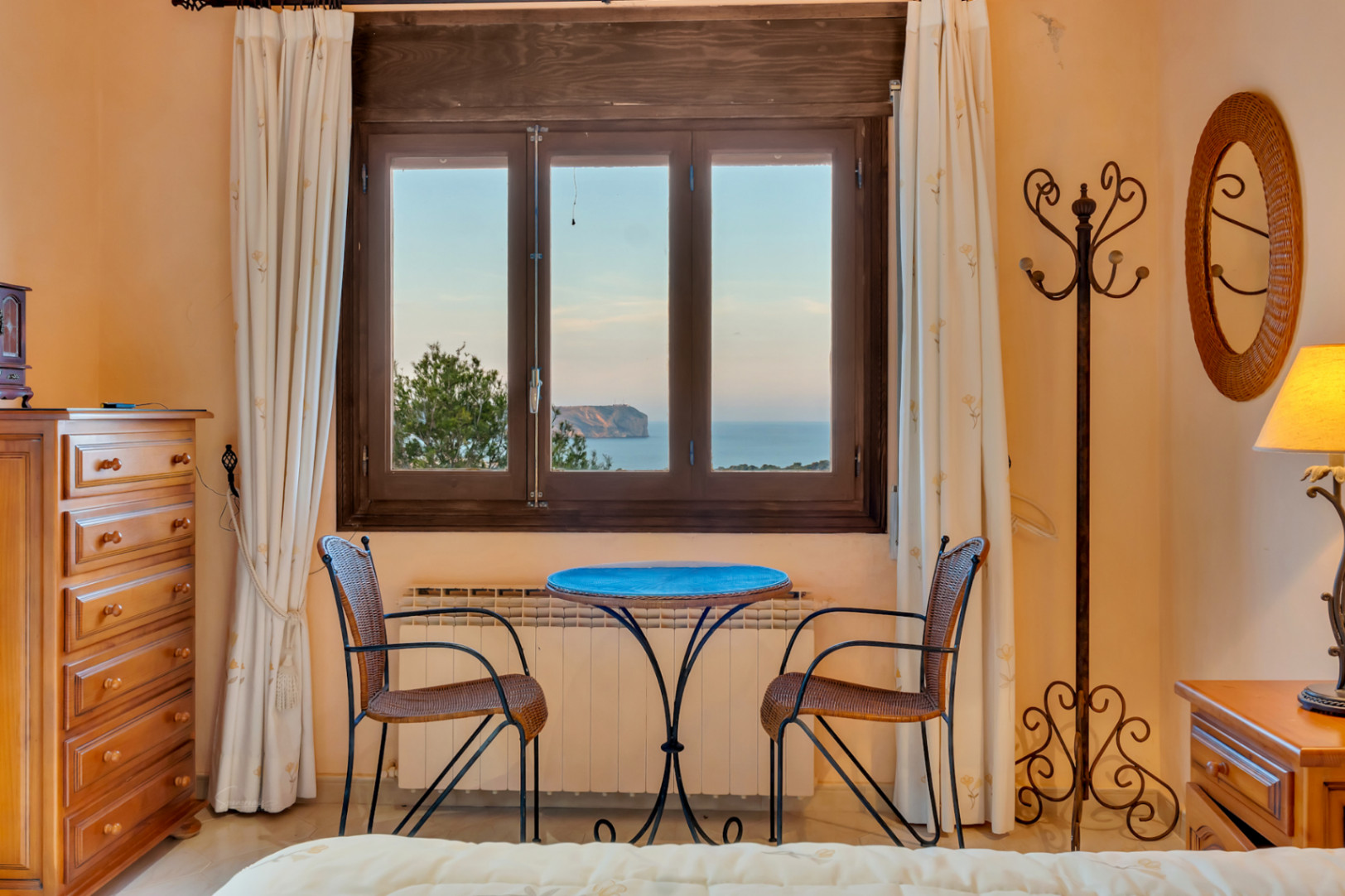 Bedroom seating area with wicker chairs and a blue table, featuring framed views of the Mediterranean Sea and Cabo San Antonio.