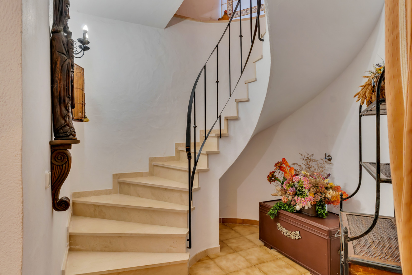 Curved interior staircase featuring light stone steps and a black wrought iron handrail. The area is finished with ceramic floor tiles and traditional decorative accents.
