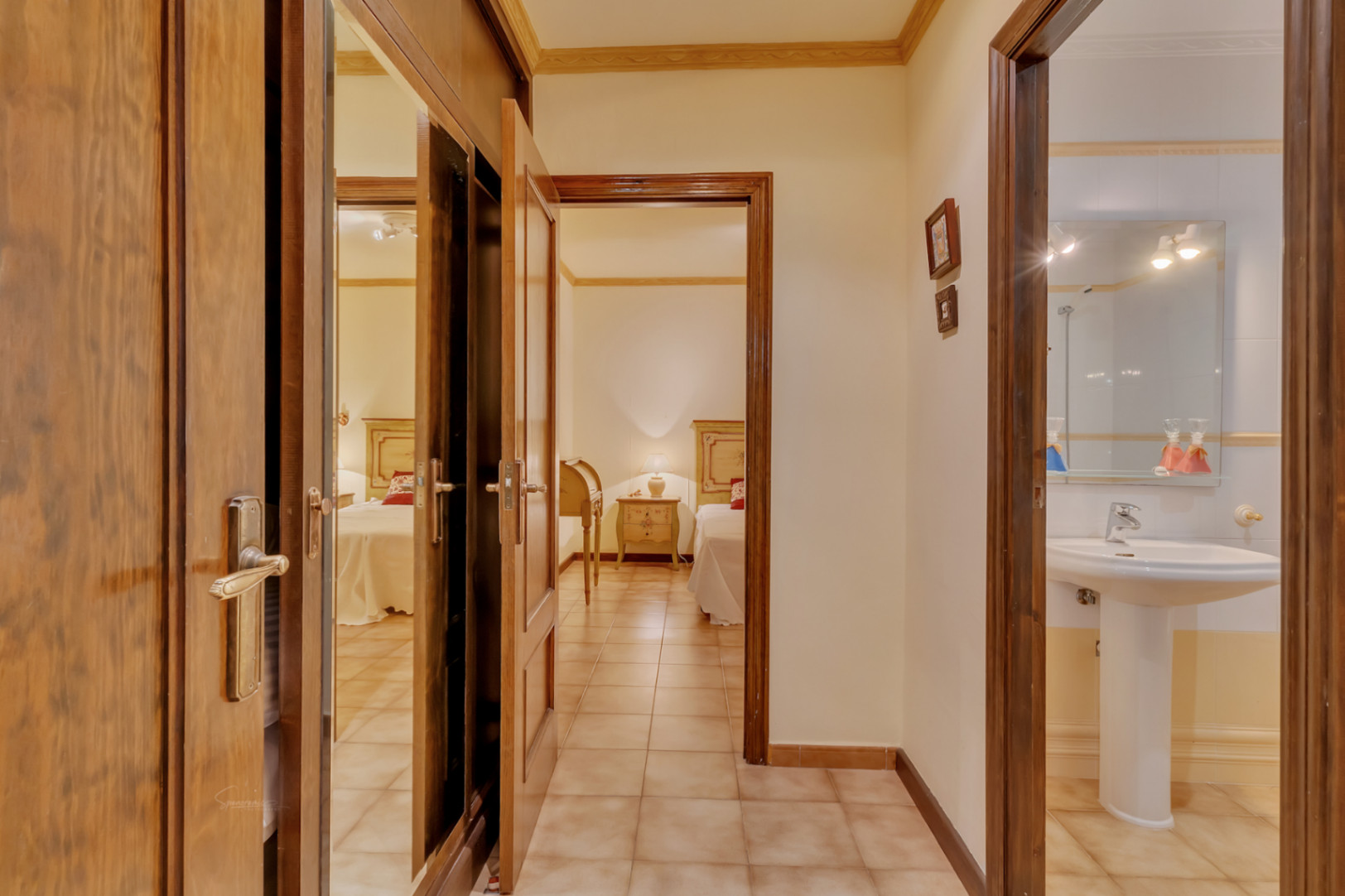Distribution hallway featuring mirrored wooden wardrobes, leading to a bedroom with twin beds and an adjacent bathroom with a pedestal sink.