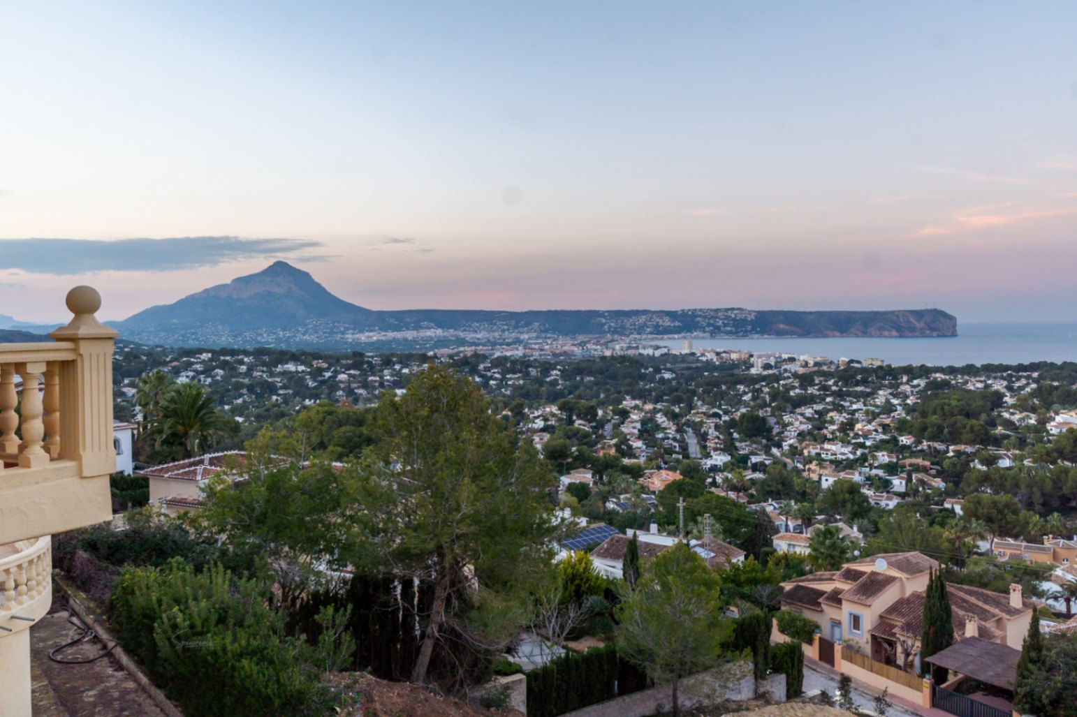 Elevated panoramic views from the terrace overlooking Jávea Bay, the Montgó mountain, and Cabo San Antonio at dusk, framed by Mediterranean greenery.