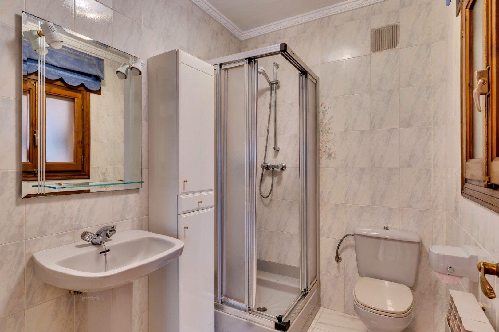 Functional bathroom featuring a pedestal sink, a corner shower cubicle with sliding doors, and a tall white storage cabinet. The walls are finished with marble-effect tiles.