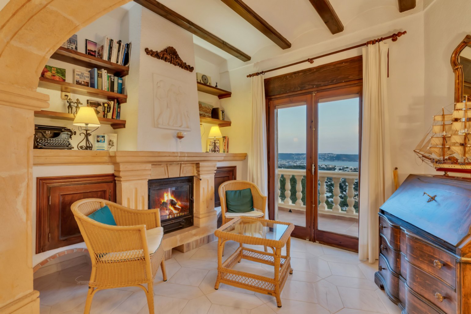 Living area featuring a stone fireplace, exposed wooden beams, and wicker seating. Large glass doors open to a terrace with panoramic bay views at dusk.