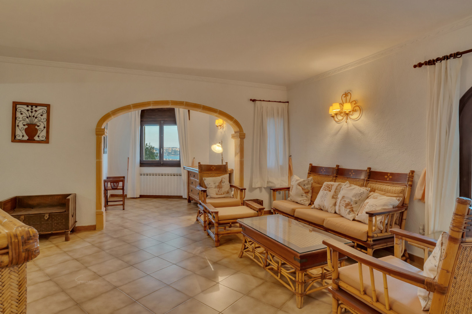 Living area featuring bamboo furniture and a traditional Tosca stone archway. The space includes ceramic flooring and a view of the bay through the adjacent room.