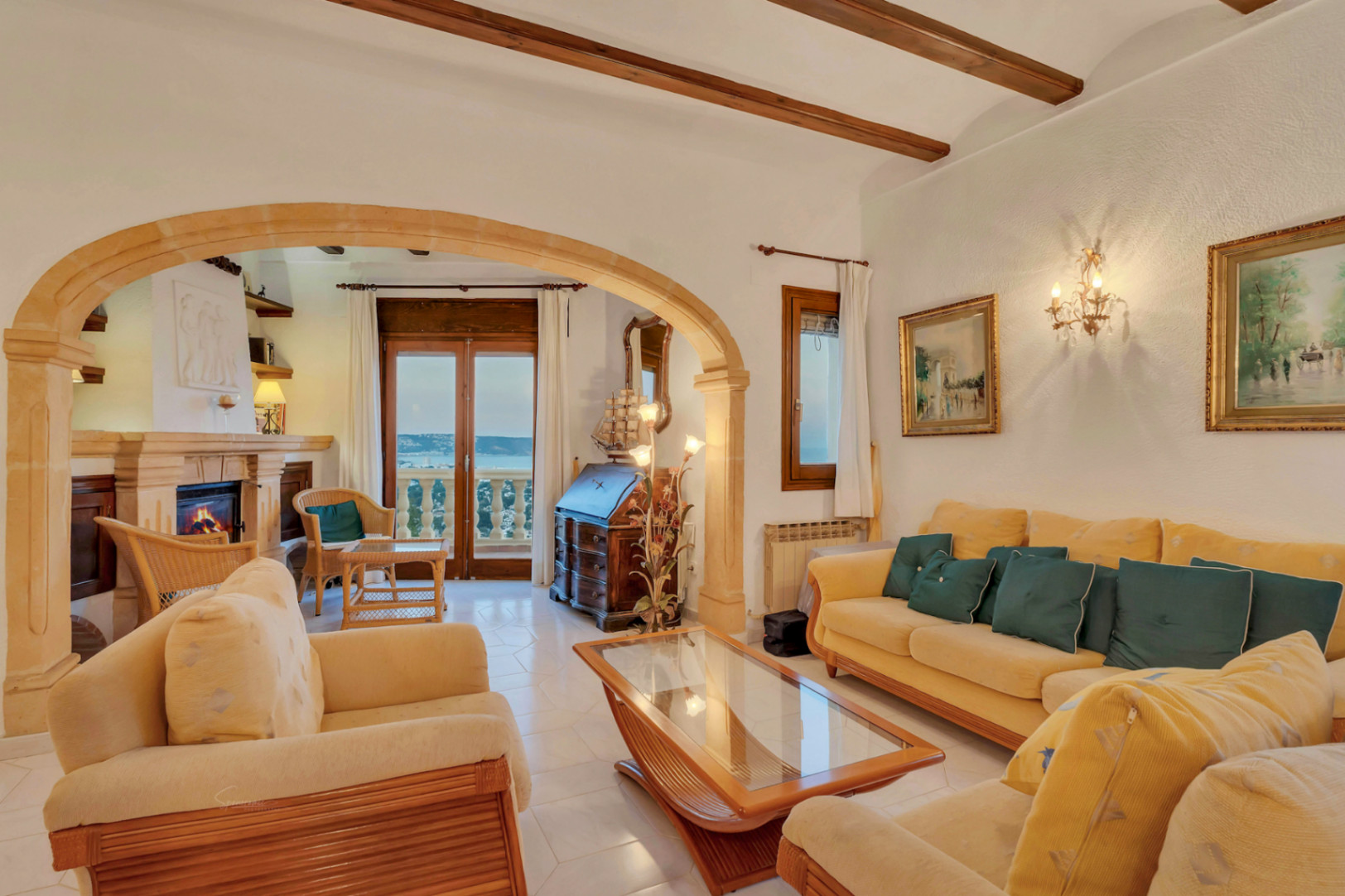 Main living area featuring exposed wooden beams, a traditional Tosca stone arch, and a fireplace. Glass doors reveal panoramic views of the bay beyond.
