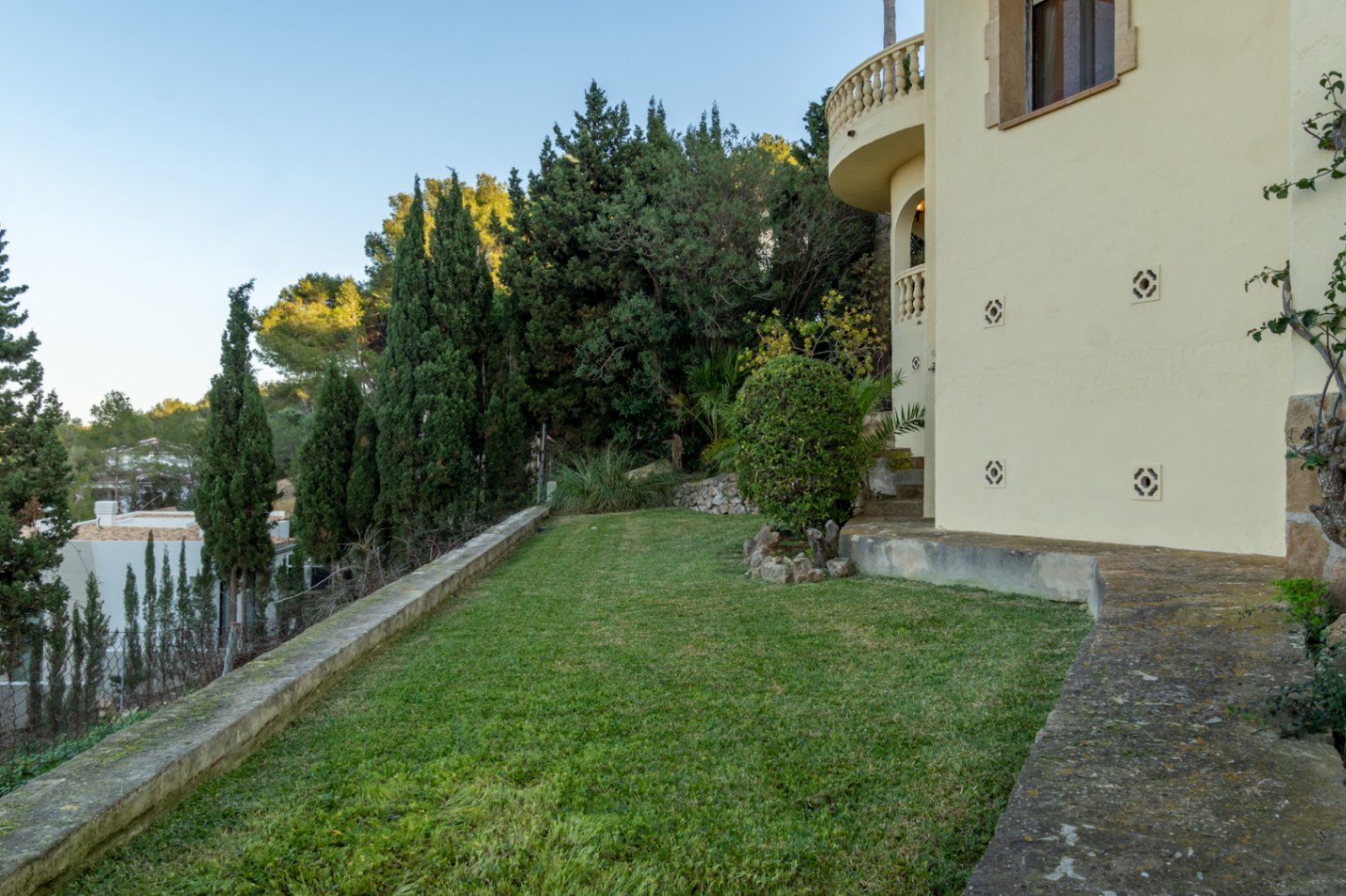Manicured Mediterranean garden featuring a level lawn, cypress trees, and stone borders. The villa's facade shows a curved balcony with traditional balustrades.