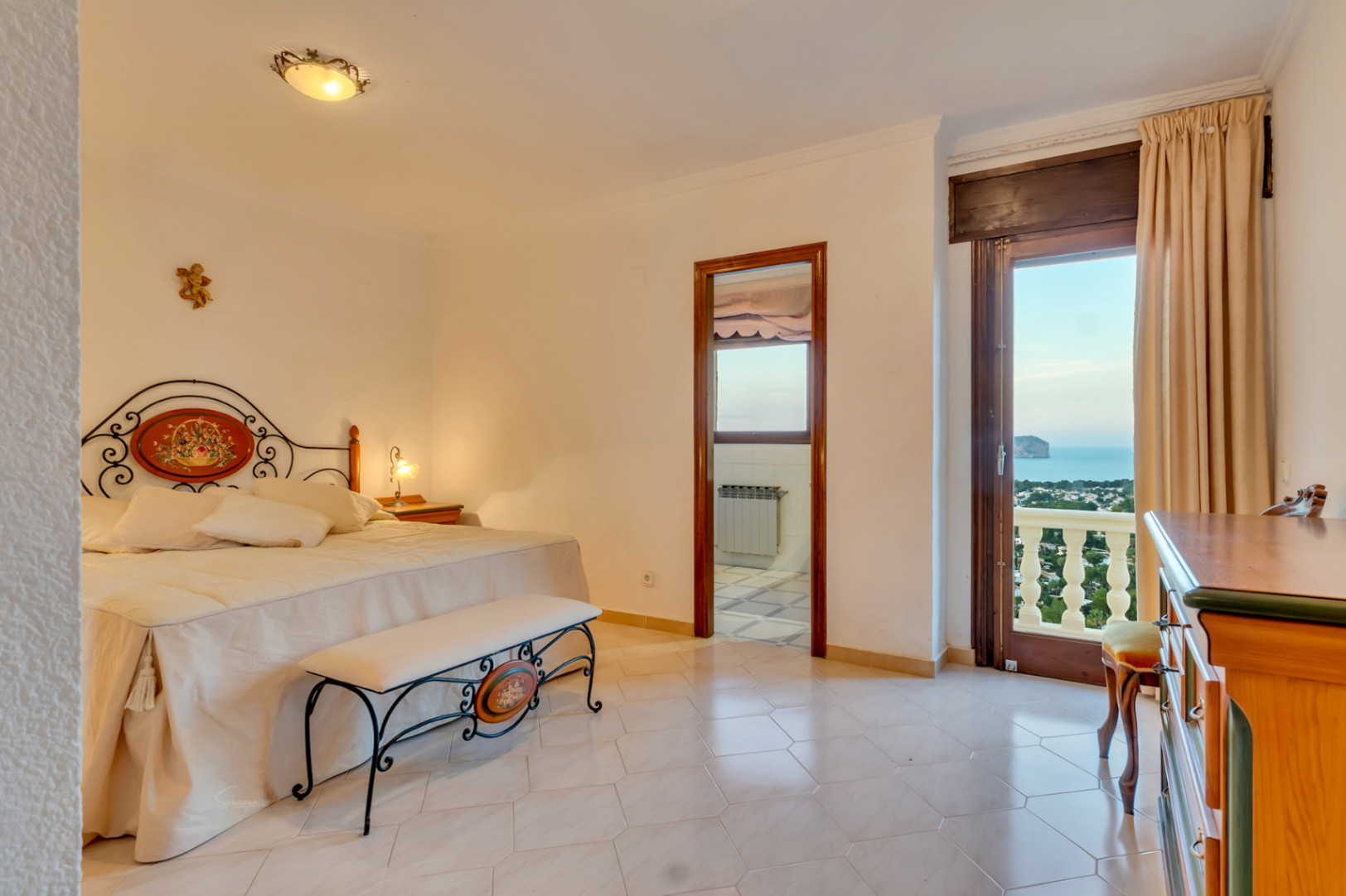 Master bedroom featuring an ornate wrought iron headboard, tiled flooring, and direct balcony access with panoramic views of the sea and Cabo San Antonio.