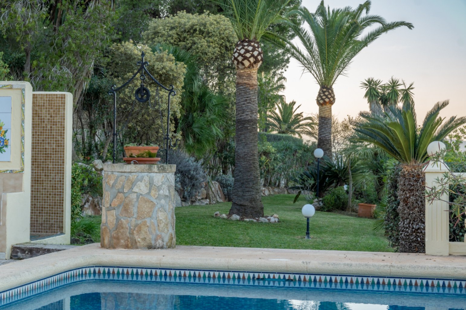 Mediterranean garden featuring a lawn, palm trees, and a stone-clad decorative well. The pool area includes an outdoor shower with mosaic tiling and globe lighting.