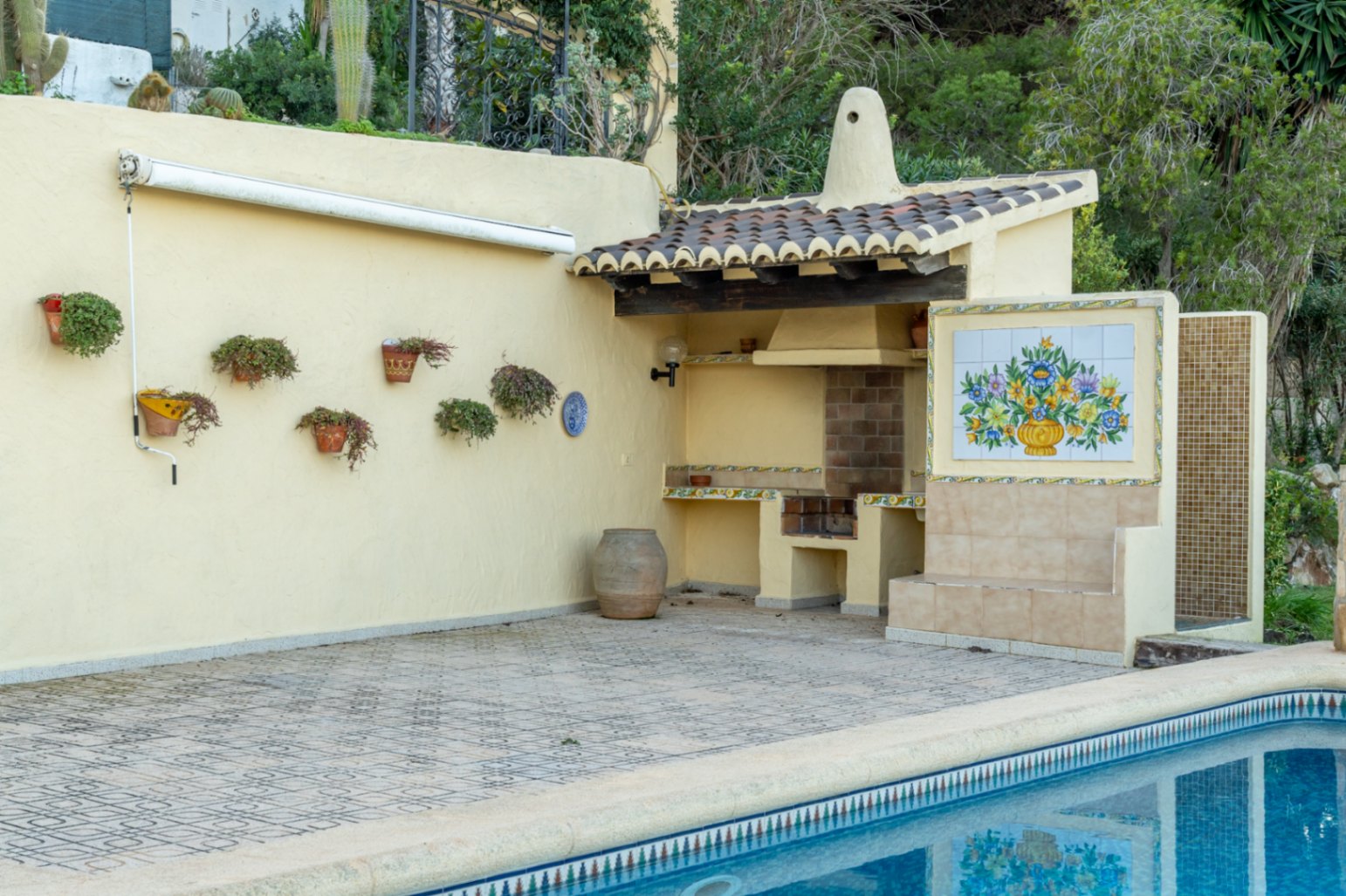 Outdoor barbecue area by the pool featuring a tiled roof, decorative floral tile mural, and a patterned stone patio with a retractable awning.