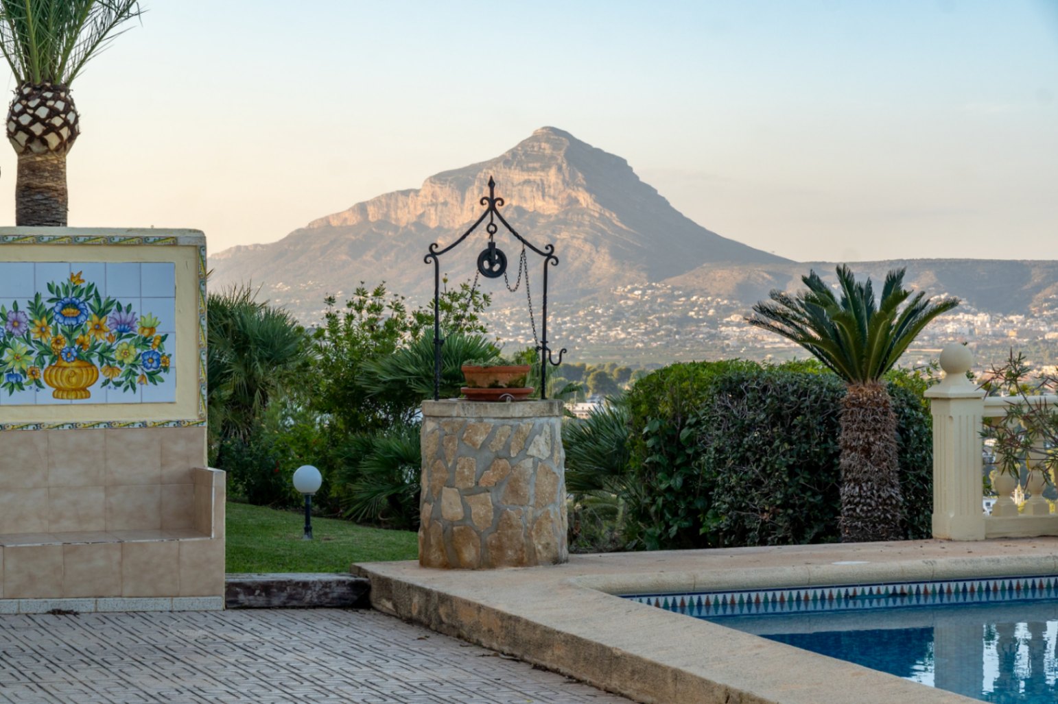 Poolside terrace featuring a decorative stone well and floral tile mural, overlooking the Montgó mountain and the Jávea coastline.