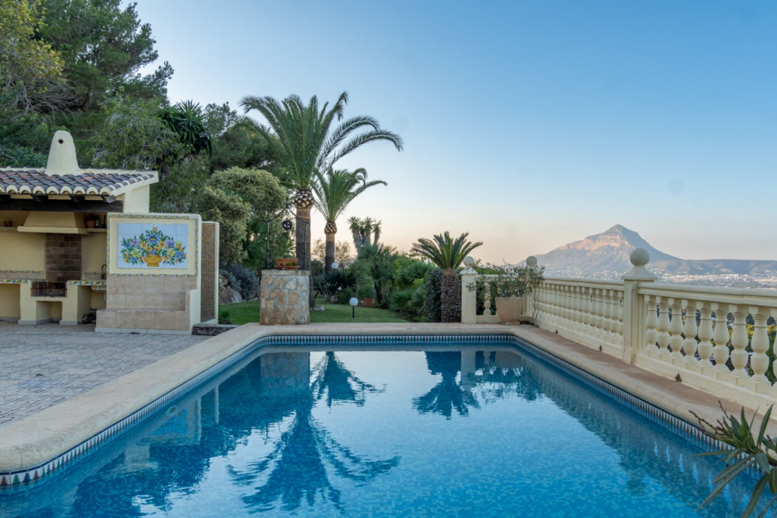 Rectangular swimming pool with blue mosaic tiles, adjacent covered barbecue area, and panoramic views of the Montgó mountain and bay across the balustrade.