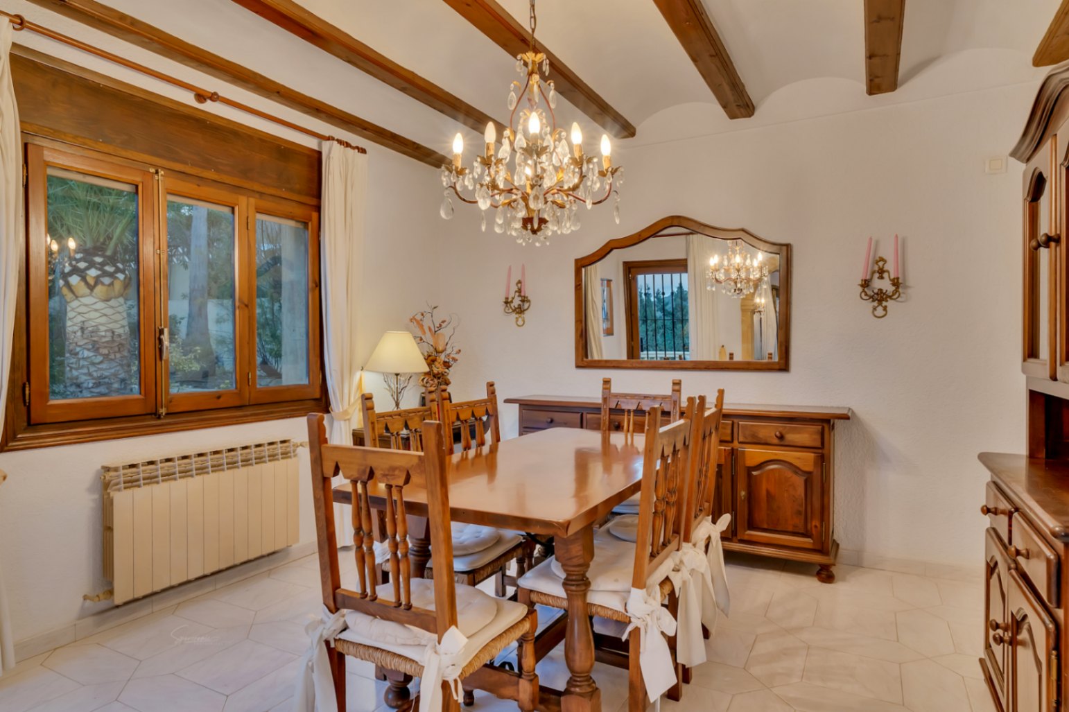 Traditional dining area showcasing exposed wooden beams, solid timber furniture, a crystal chandelier, and light-colored geometric floor tiling.