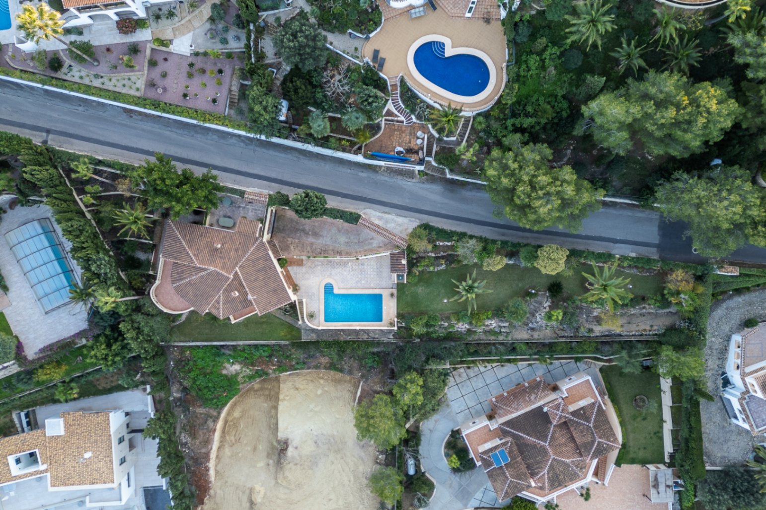 Zenithal aerial view of the property showcasing the plot layout, L-shaped swimming pool, paved terraces, and Mediterranean garden with mature palm trees.