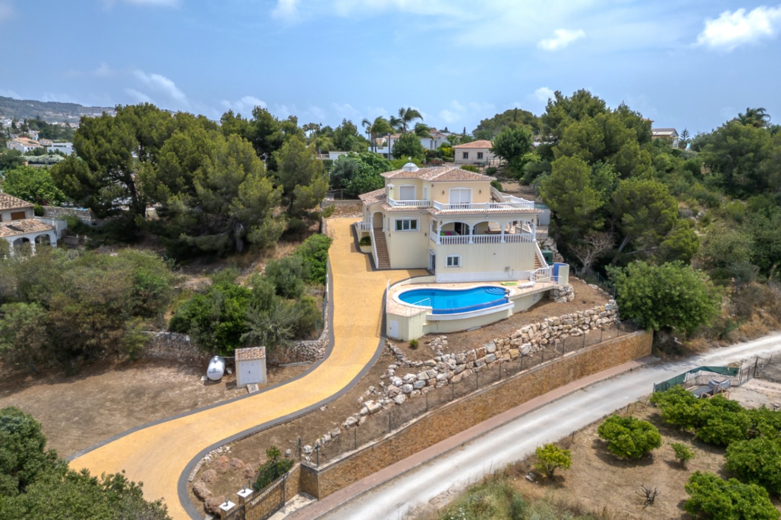 Aerial view of the elevated villa featuring a curved infinity pool, expansive terraces, and a long paved driveway set within Mediterranean pine forests.