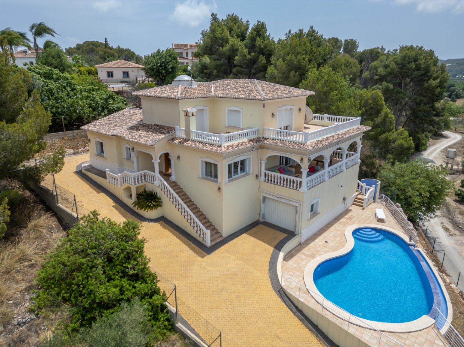 Aerial view of the villa featuring a cream facade, arched naya, and a curved pool, surrounded by expansive white balustraded terraces and lush pine trees.