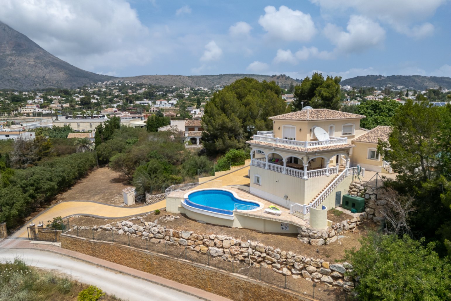 Aerial view of the villa featuring a curved infinity pool, arched terraces with white balustrades, and panoramic views of the Montgó mountain and valley.