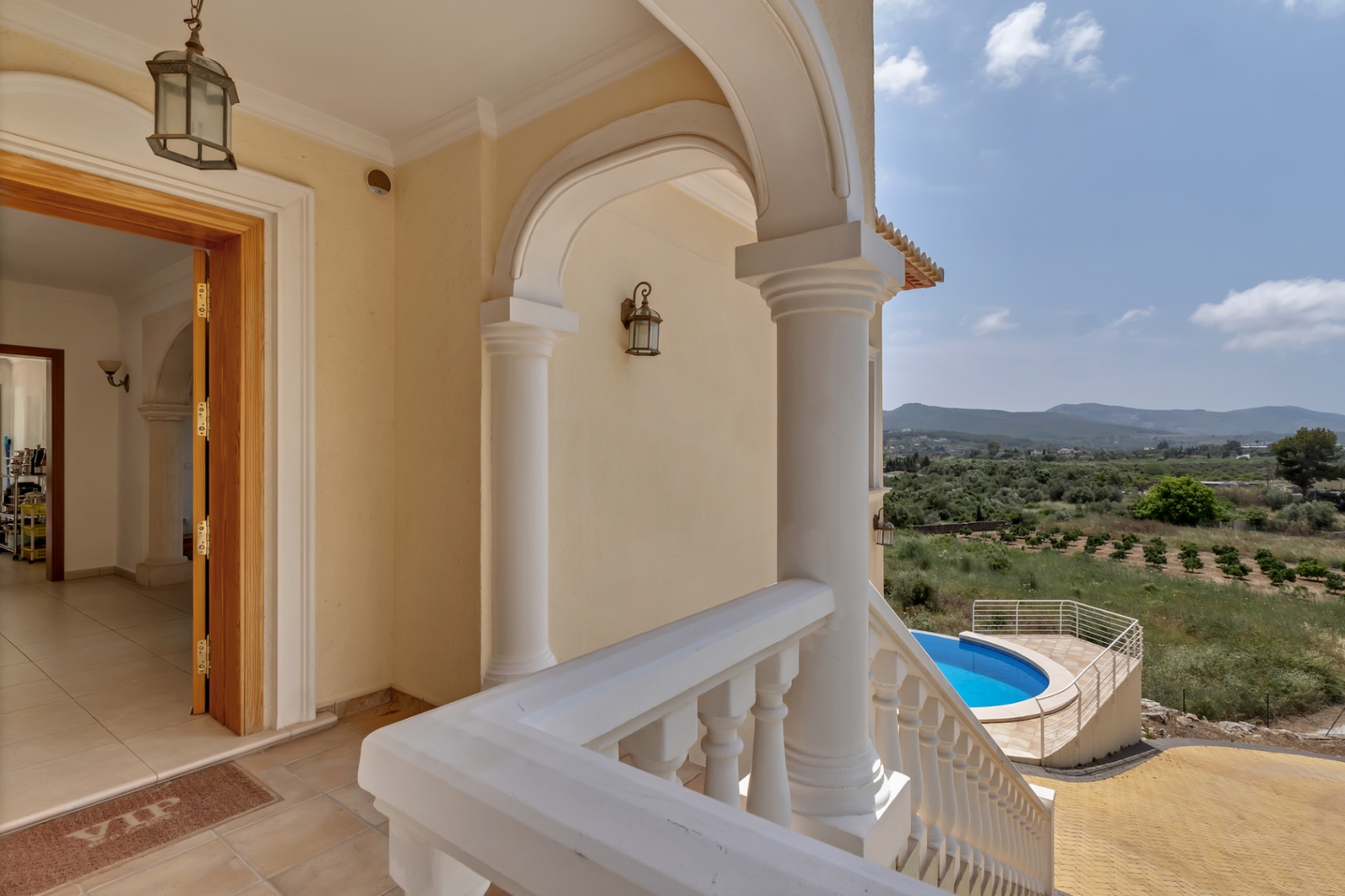 Arched entrance porch with white columns and balustrade, overlooking the circular pool and valley. Features terracotta tiling and a solid wood front door.