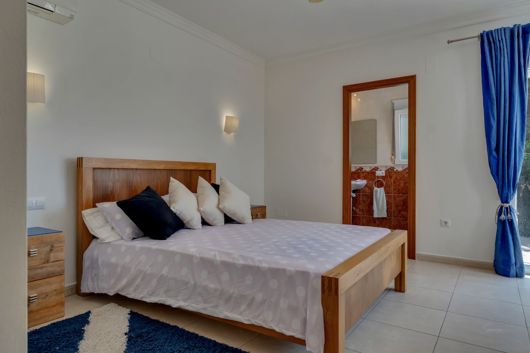 Bedroom featuring a wooden bed frame, tiled floors, and an en-suite bathroom with reddish tiles. Includes wall-mounted lighting and split-system air conditioning.