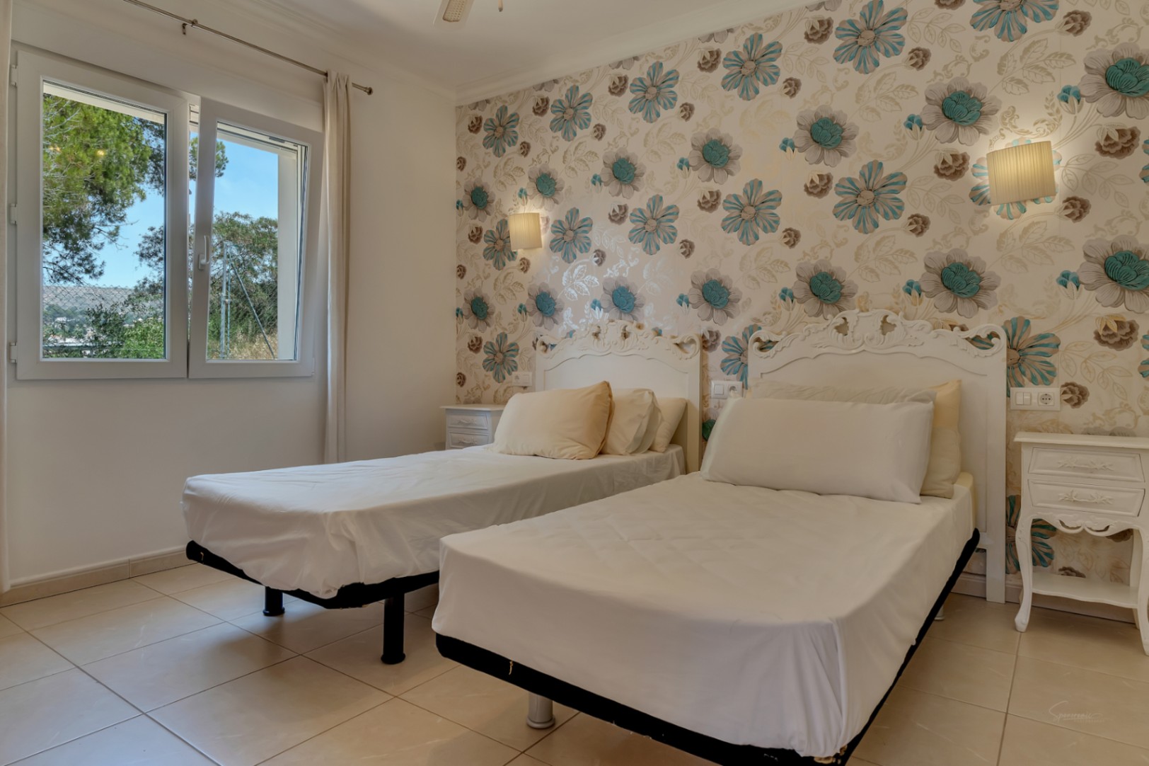 Bedroom featuring twin beds with ornate white headboards and floral patterned wallpaper. Includes light tiled flooring and a window overlooking the Mediterranean greenery.
