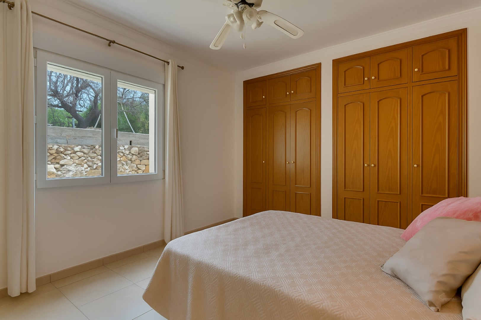 Bright bedroom featuring built-in wooden wardrobes, a ceiling fan, and a window overlooking the garden's natural stone wall.