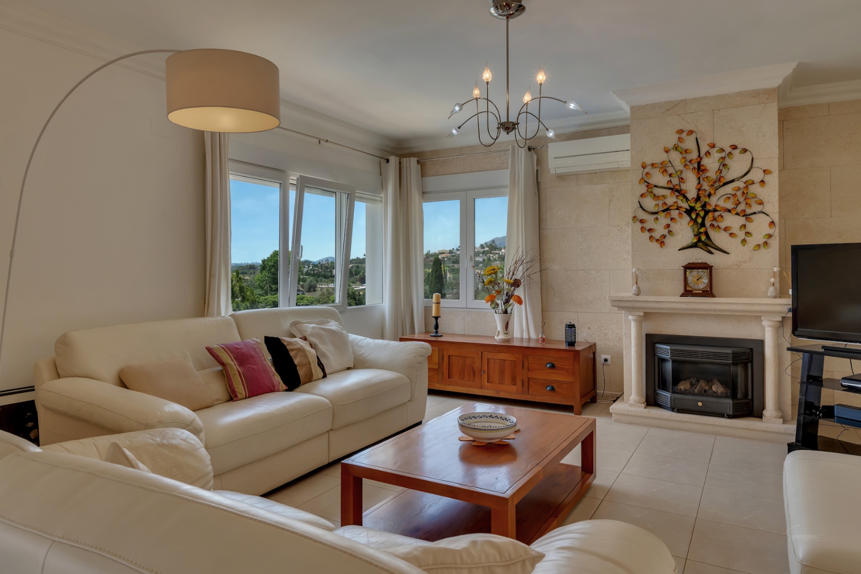 Bright living room featuring white leather sofas, a stone-clad fireplace with wood-burning stove, and large windows showcasing panoramic valley views.
