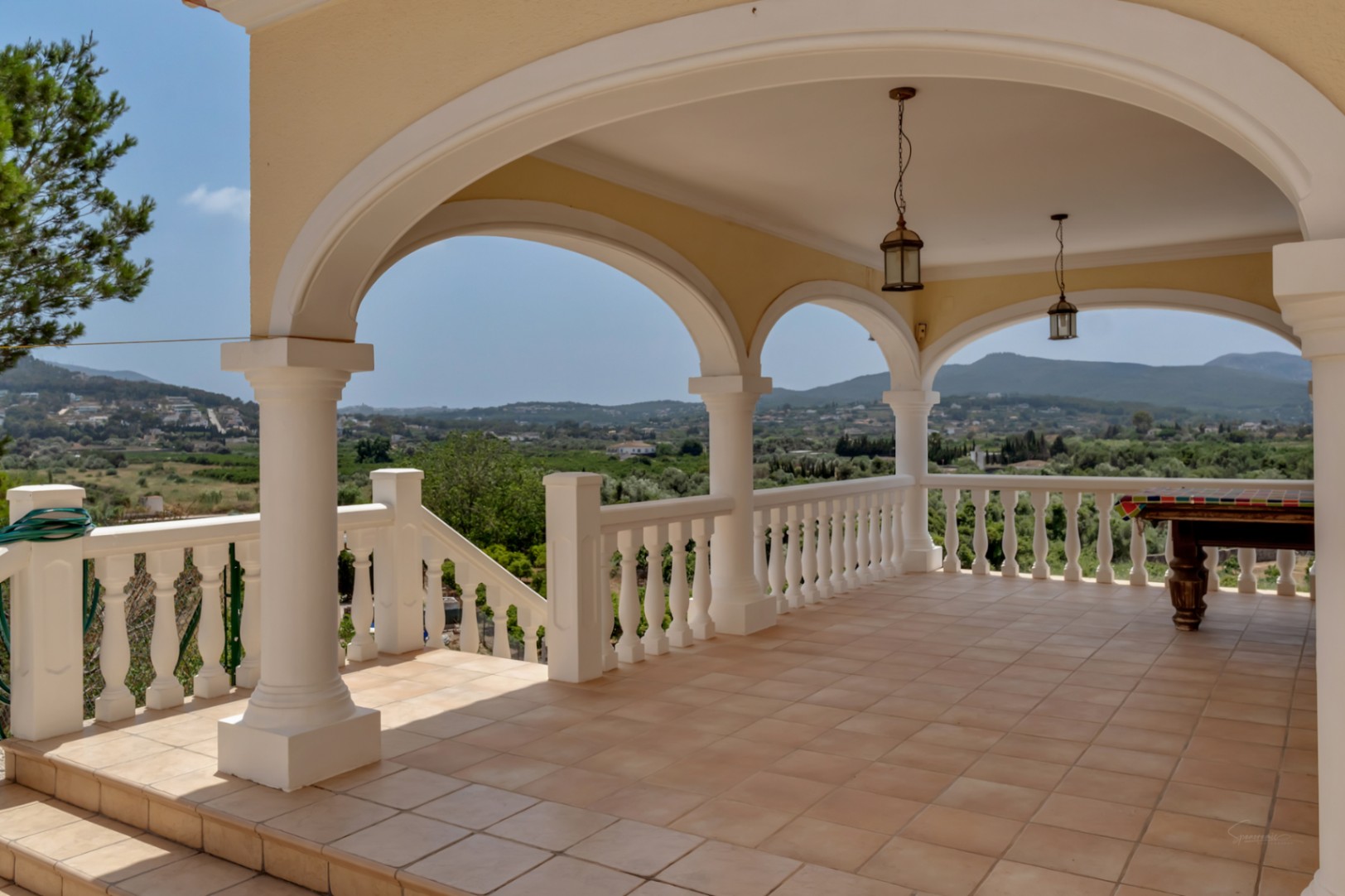 Covered arched terrace with white columns and balustrade railing. Features terracotta tiling, lantern pendants, a pool table, and panoramic valley views.