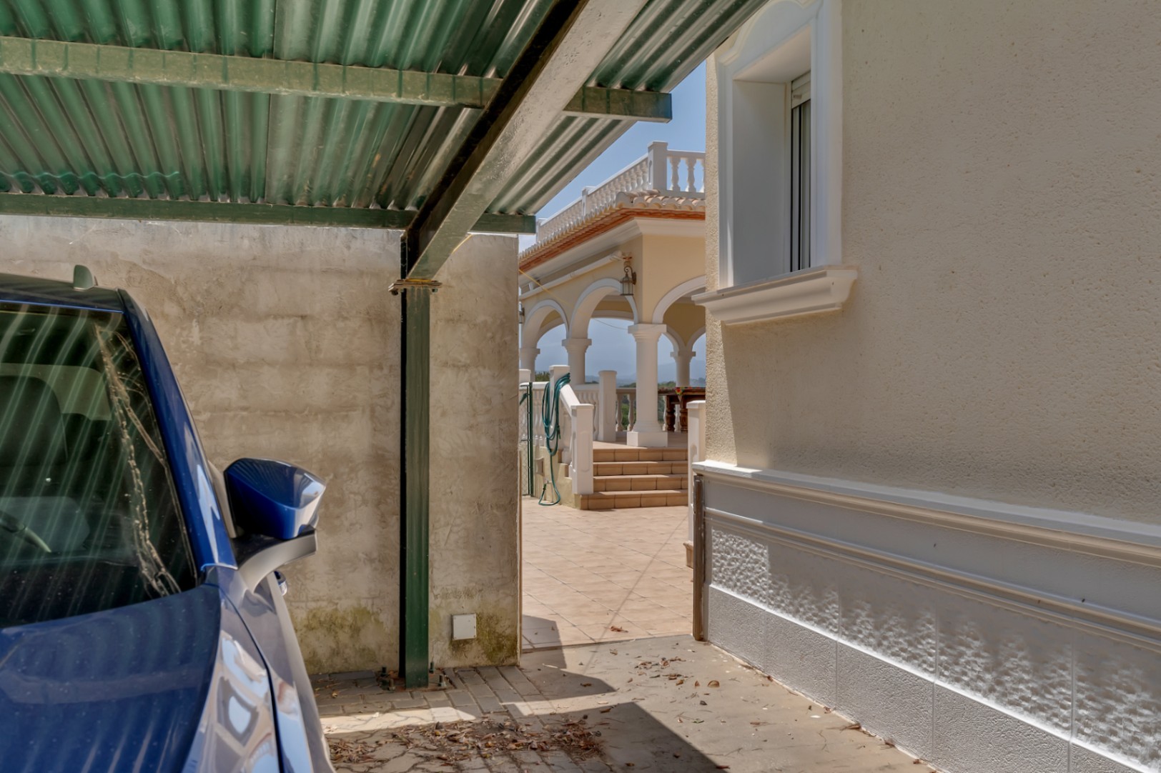 Covered parking area with a metal carport adjacent to the main villa, featuring a paved path leading to the elevated arched terrace and naya.