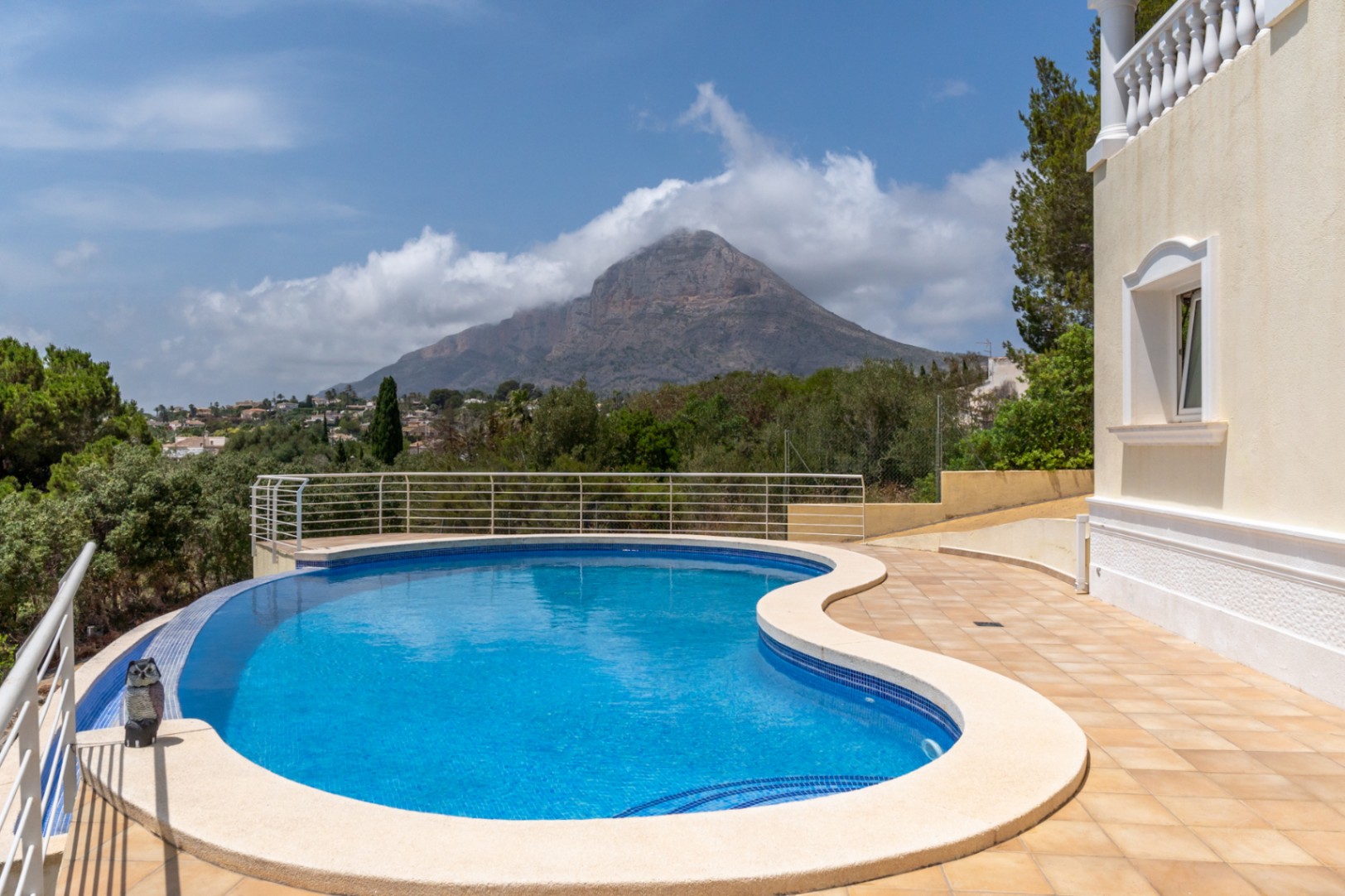 Curved infinity pool featuring blue mosaic tiling and panoramic views of the Montgó mountain, set within a spacious tiled terrace with metal railings.