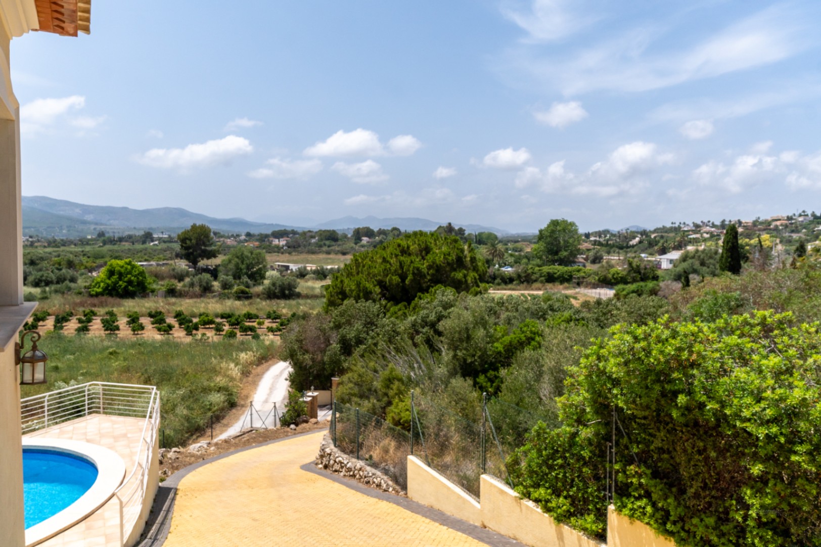 Elevated view showing the paved driveway, a curved swimming pool with white railings, and panoramic vistas across the lush Jávea valley and orange groves.