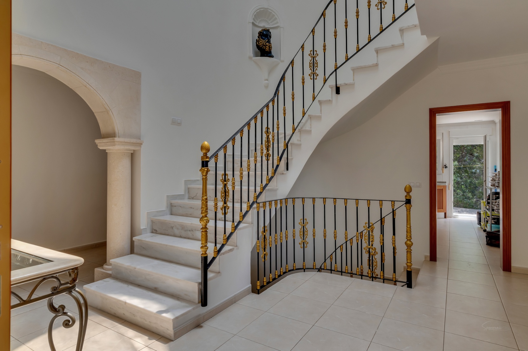 Entrance hall featuring a marble staircase with ornate gold and black wrought iron railings, adjacent to a carved stone archway and column.