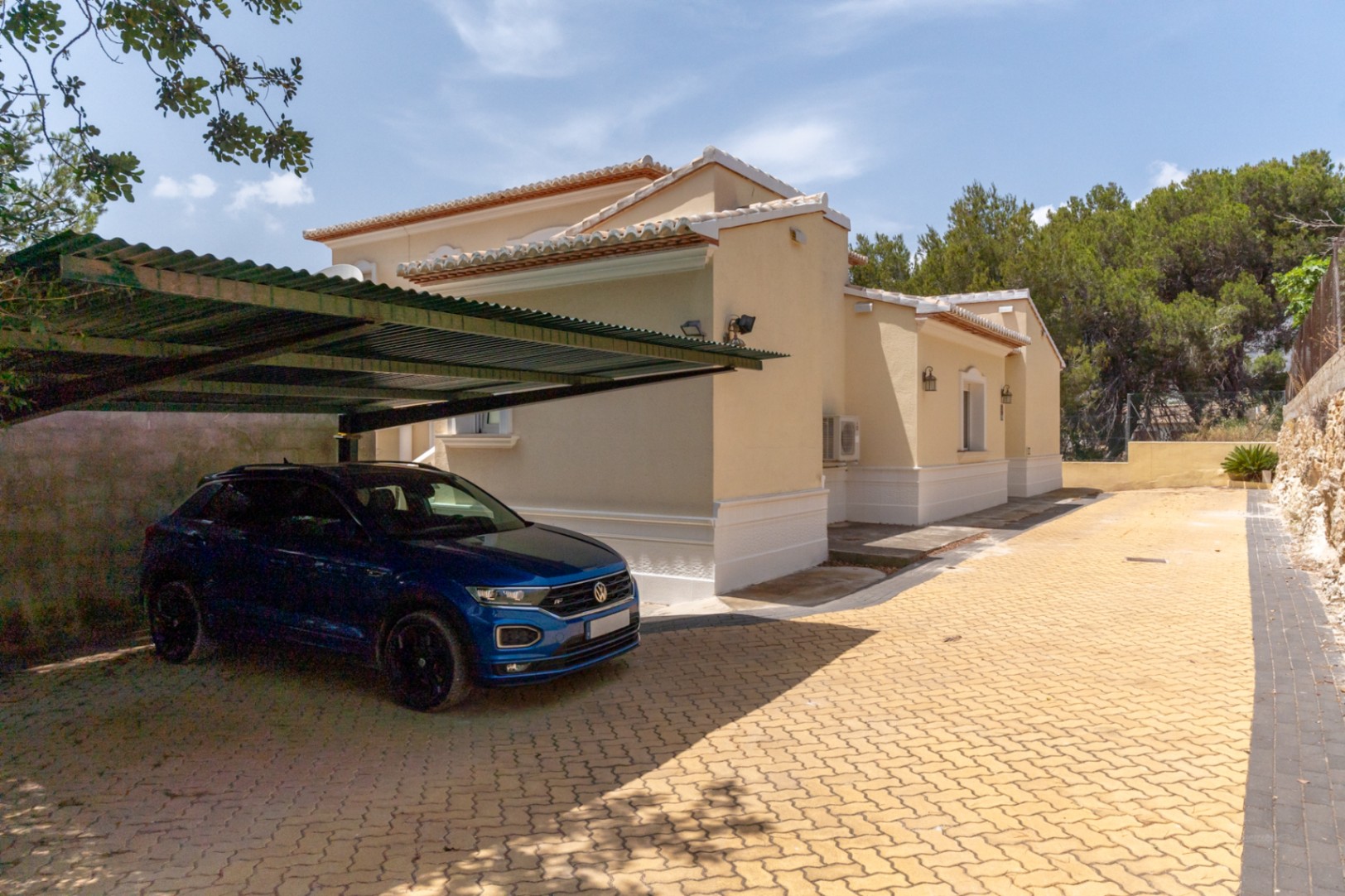 Exterior parking area featuring yellow interlocking pavers and a covered carport structure adjacent to the villa's cream-colored side facade.