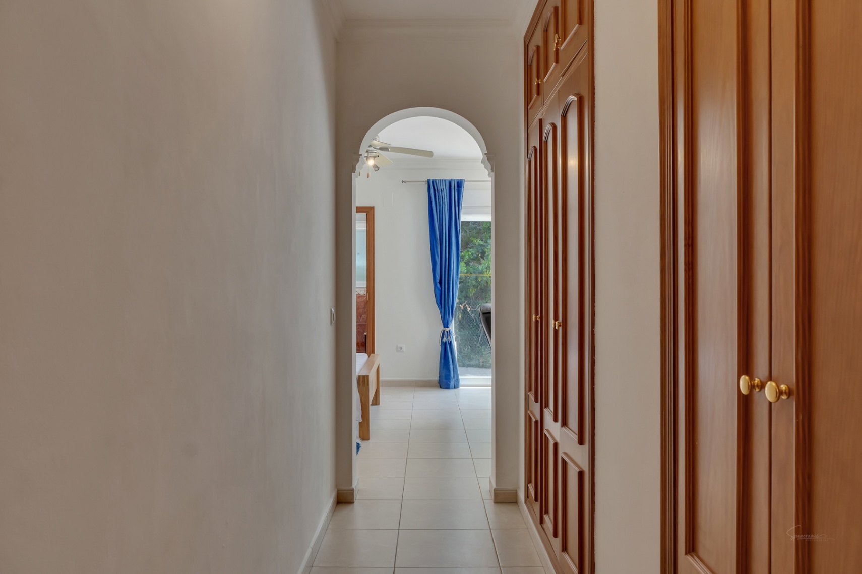 Hallway with large built-in wooden wardrobes and ceramic flooring, connecting via an archway to a bright bedroom featuring a ceiling fan and blue curtains.