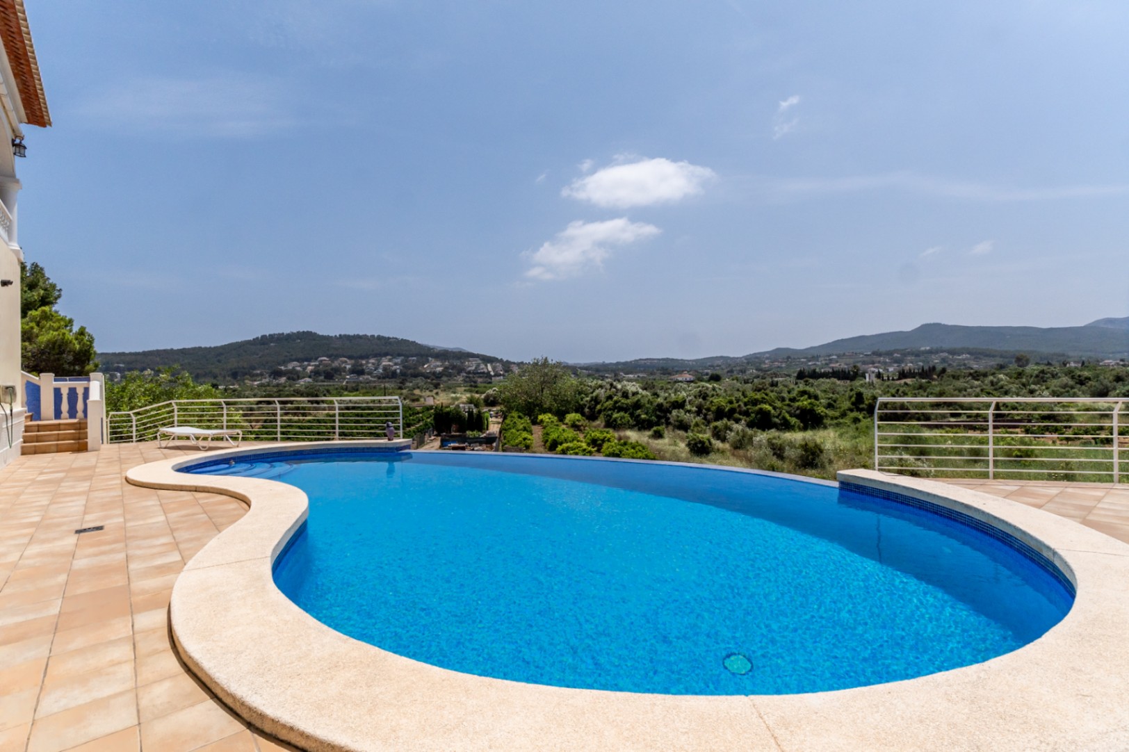 Infinity pool with a curved design offering panoramic views of the Jávea valley, featuring a spacious tiled terrace and white metal railings.