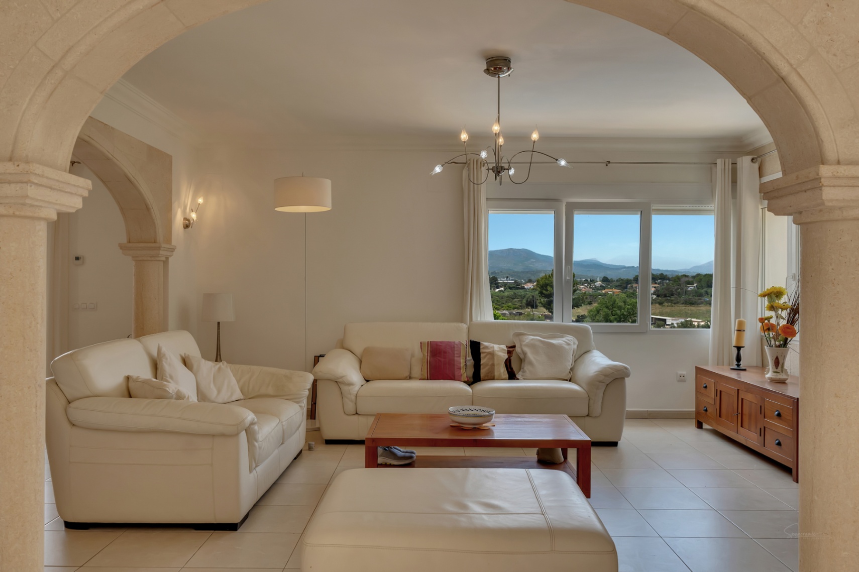 Living room featuring white leather sofas framed by traditional stone arches, offering panoramic valley and mountain views through a large window.
