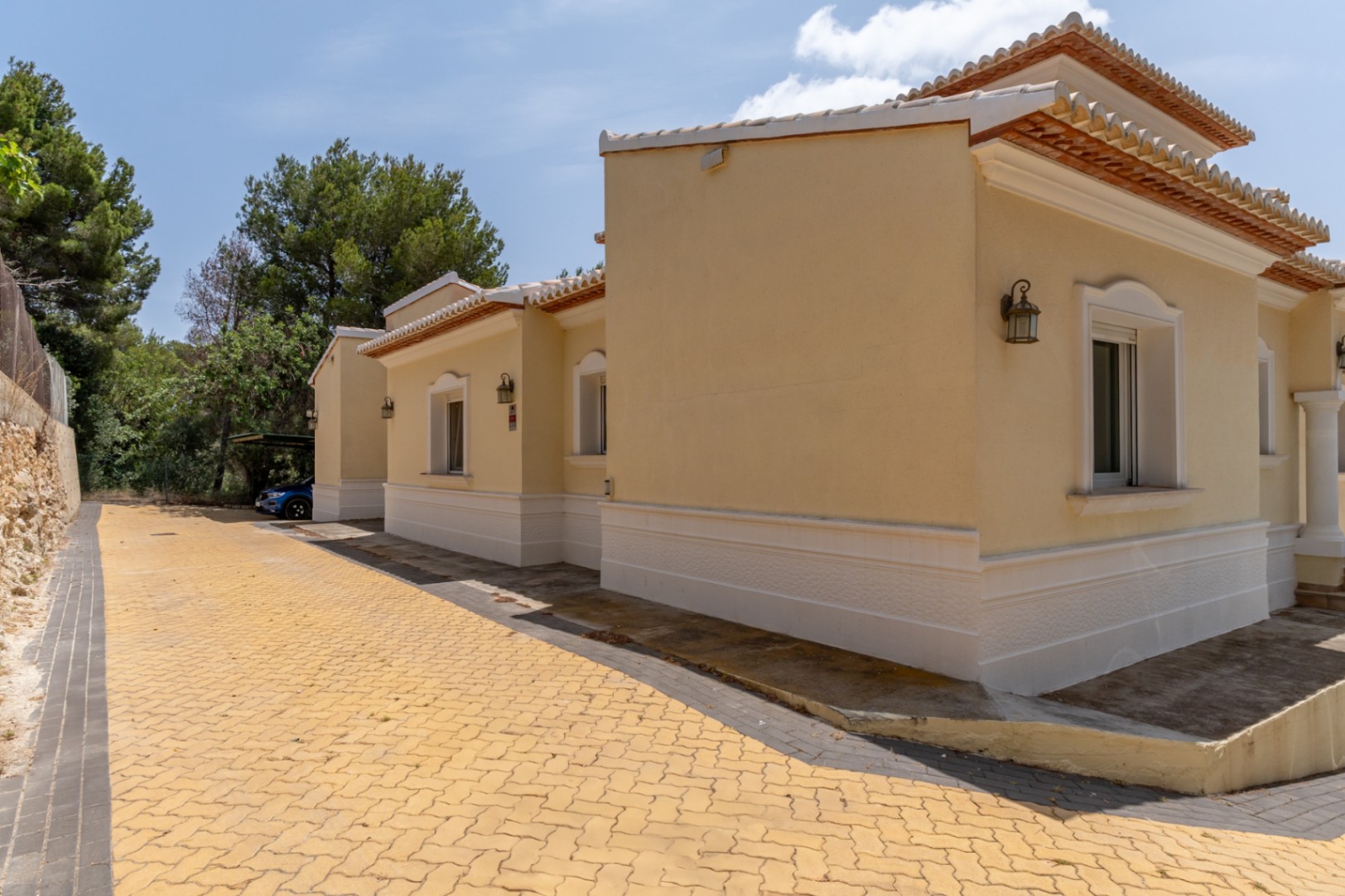 Ochre exterior facade with white moldings, featuring a paved driveway leading to a covered parking area surrounded by Mediterranean pine trees.
