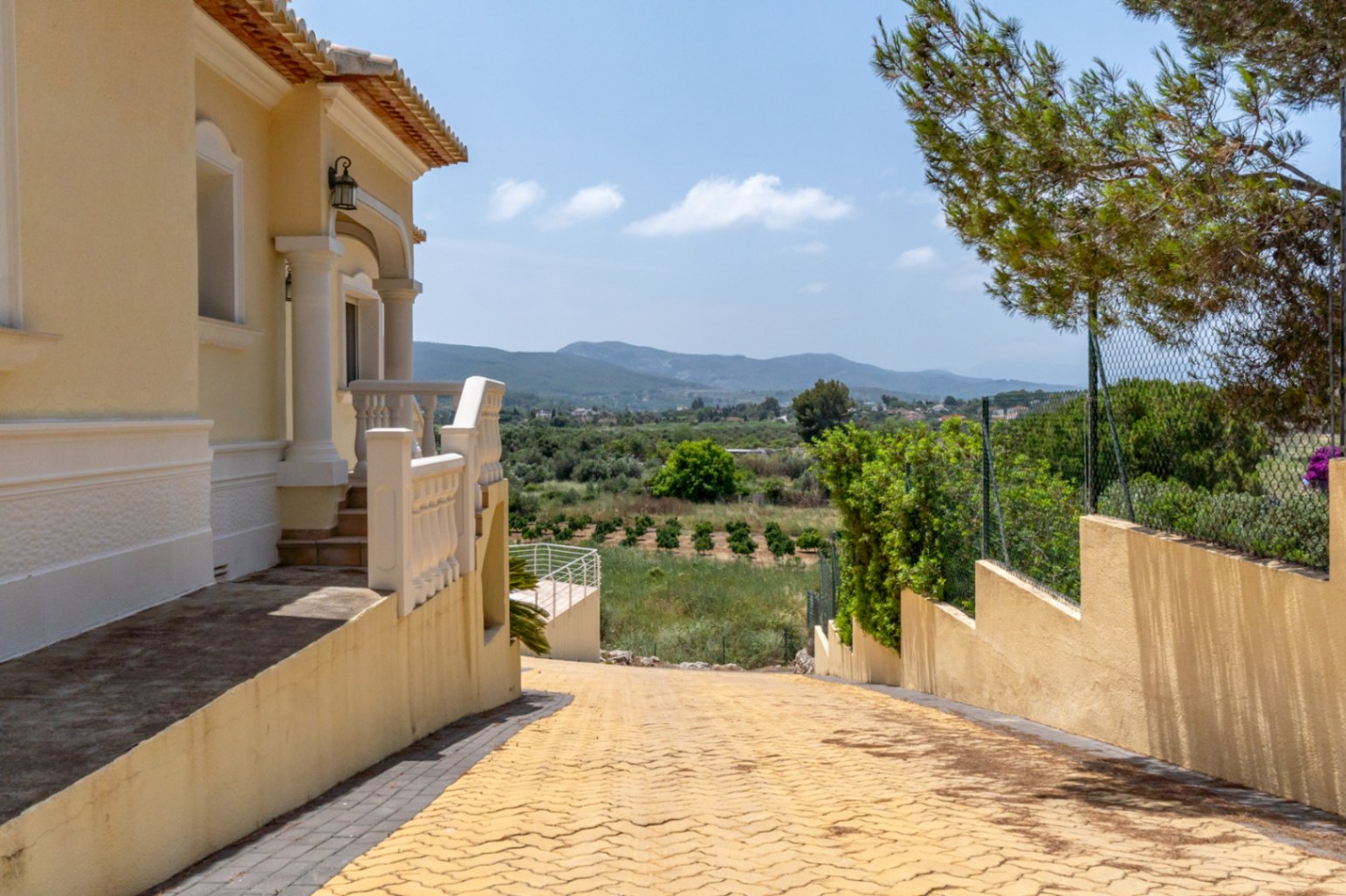 Paved driveway descending past the villa's side entrance, featuring classic white balustrades and open views of the Mediterranean valley and mountains.