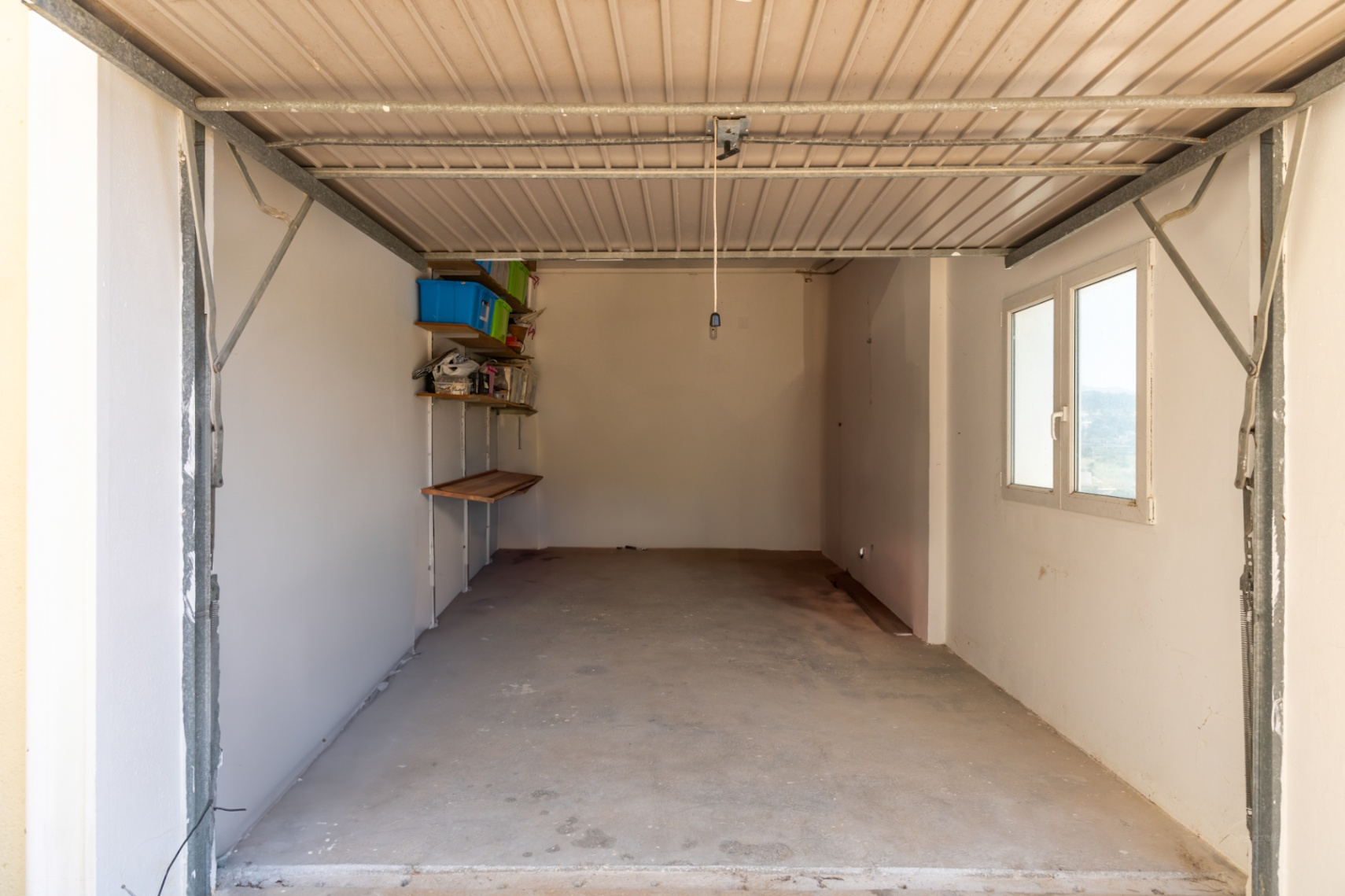 Private garage featuring a concrete floor, white walls, and a side window for natural light. Equipped with wall-mounted shelving and a metal up-and-over door.