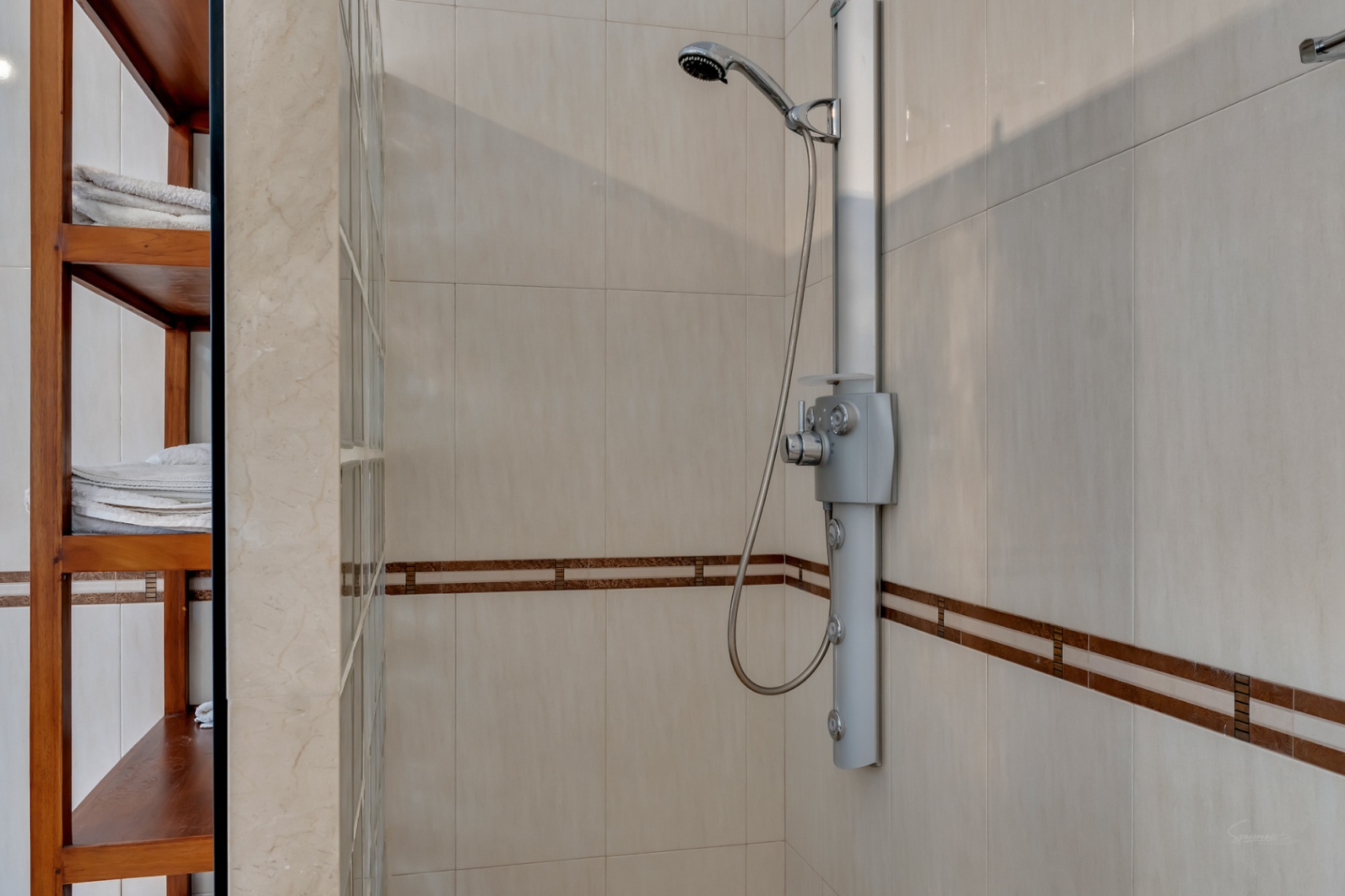Shower area featuring a hydromassage column, neutral cream tiling with a decorative border, and built-in wooden shelving adjacent to a glass block wall.
