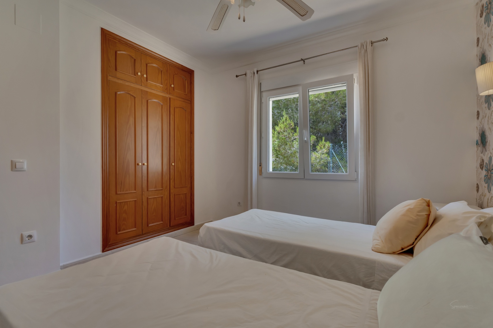 Twin bedroom featuring built-in wooden wardrobes, a ceiling fan, and a window overlooking the surrounding Mediterranean trees.