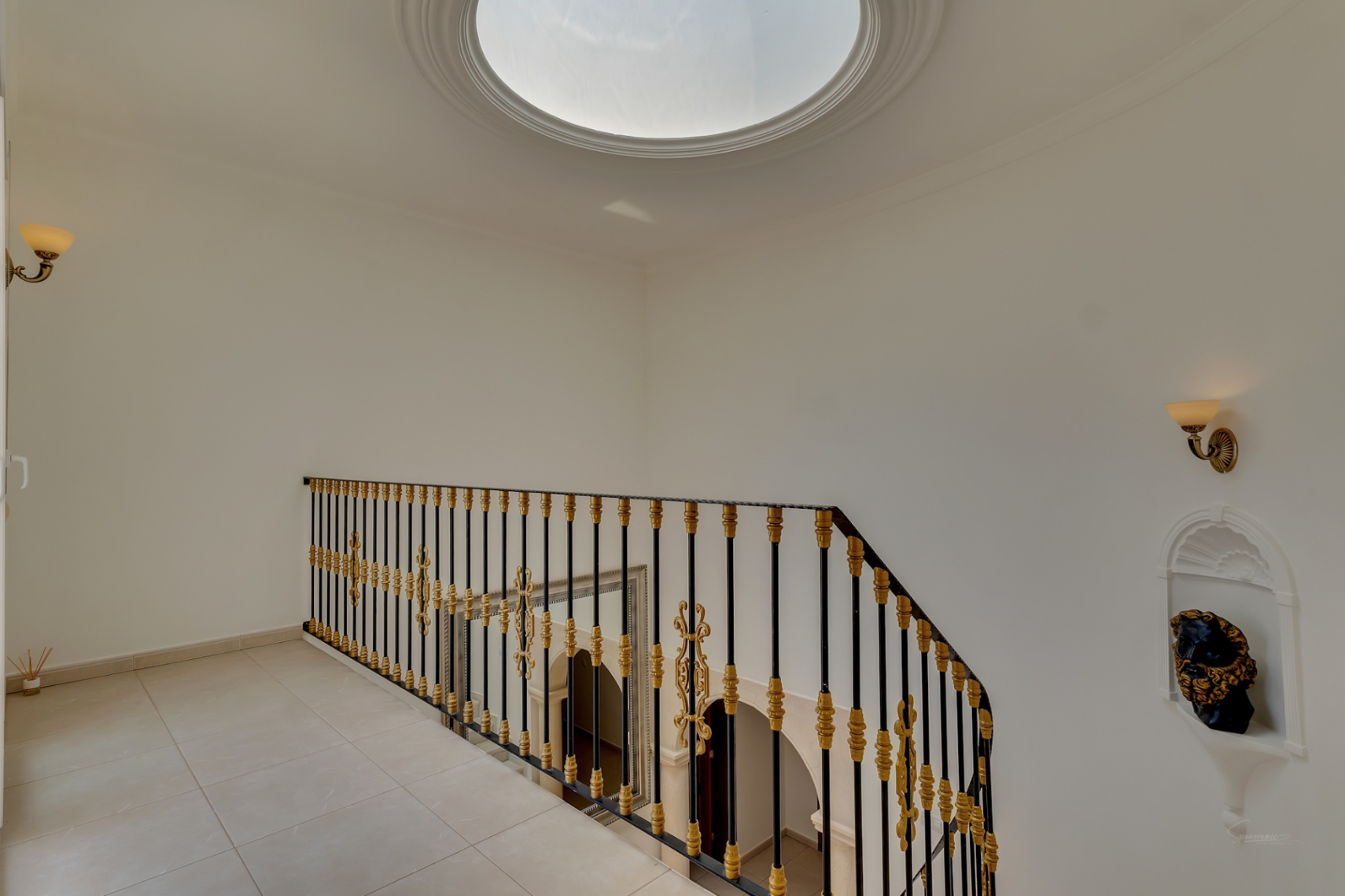 Upper landing featuring a circular skylight, wrought iron railing with gold accents, and a decorative wall niche. Overlooks the double-height entrance hall.