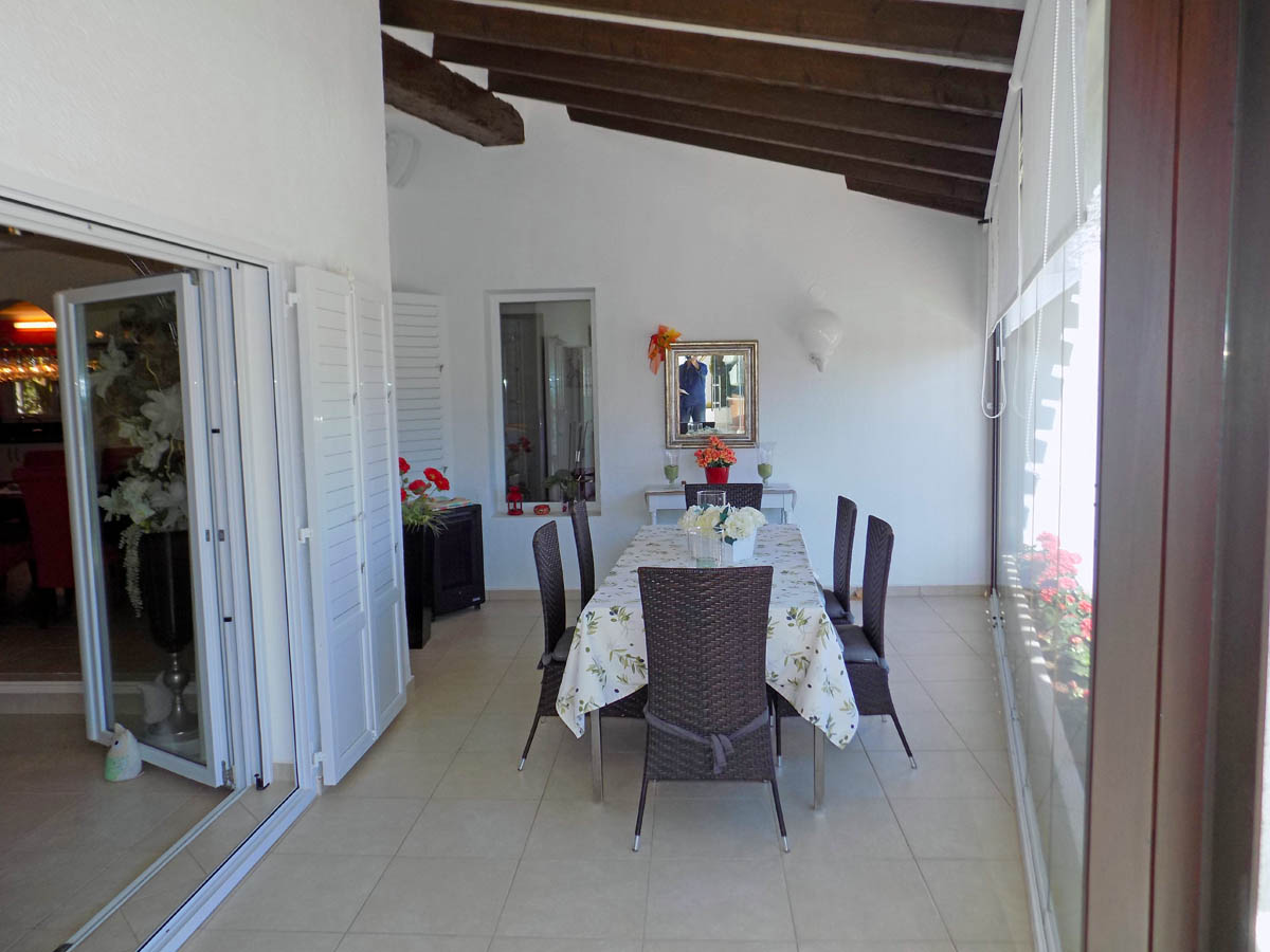 Enclosed terrace featuring exposed wooden ceiling beams, a dining area with wicker chairs, and large glass windows providing ample natural light.