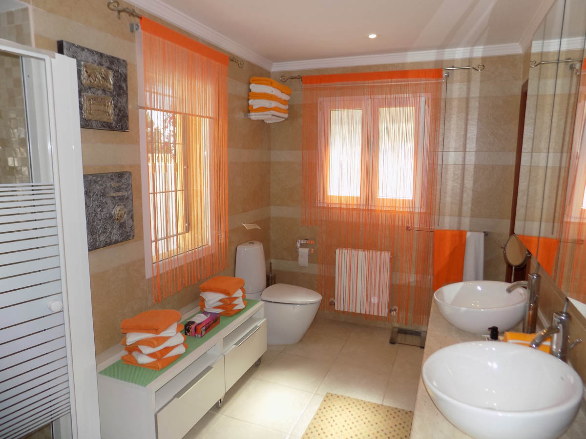 Full bathroom featuring double vessel sinks, cream-toned tiled walls, and natural light from two windows. It is equipped with a radiator and modern white cabinetry.