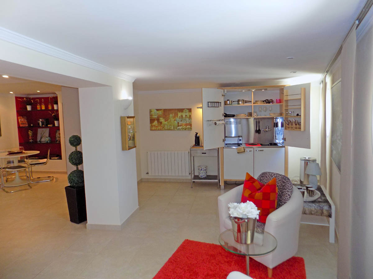 Lower-level living area featuring a compact integrated kitchenette, a seating zone with a white armchair and red rug, and a dining space with built-in shelving.