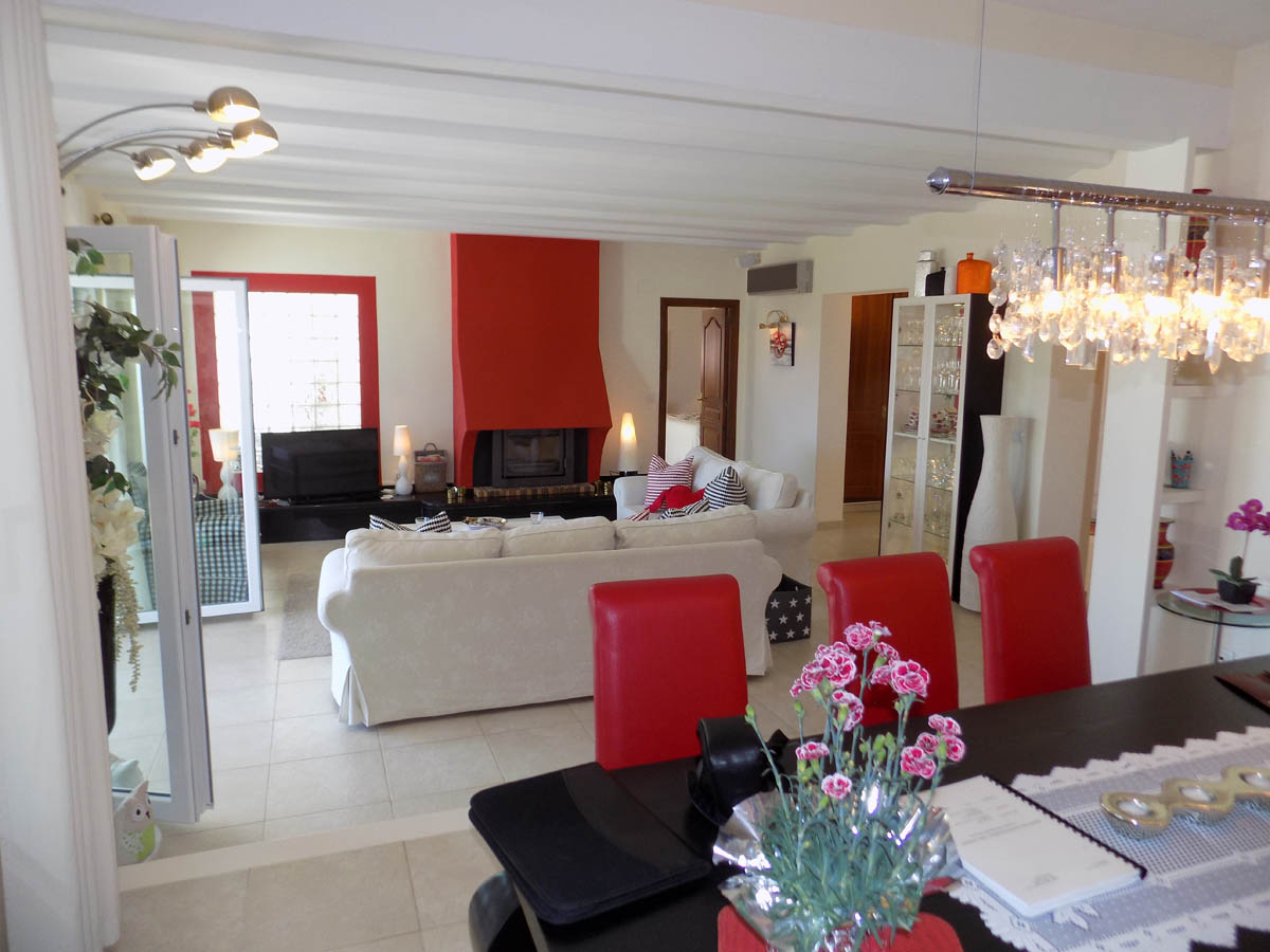 Open-plan living and dining area featuring white beamed ceilings, a bold red fireplace, and bright tiled floors with access to the terrace.